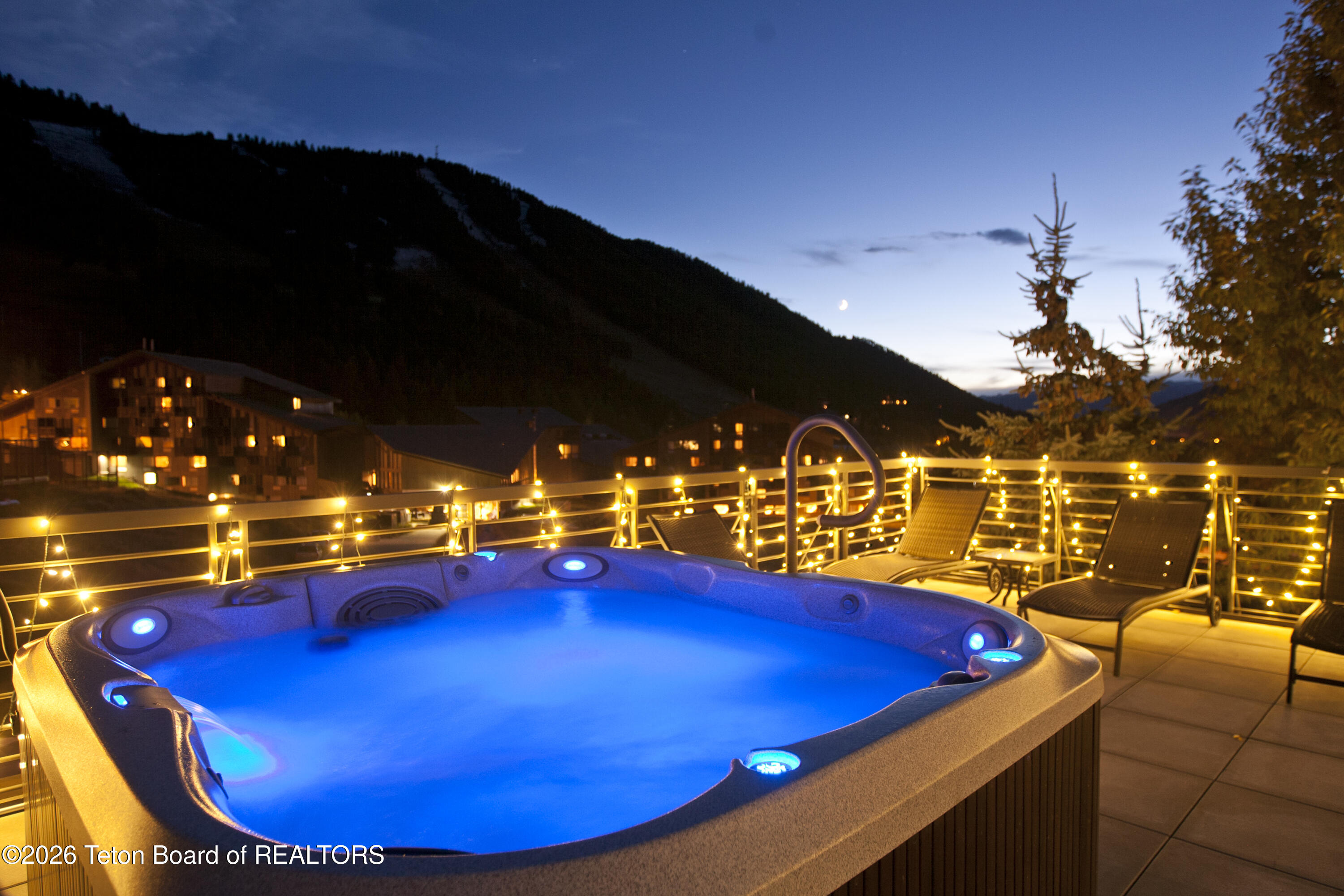 Outdoor hot tub glowing blue on a terrace strung with lights, with a mountain slope in the background.