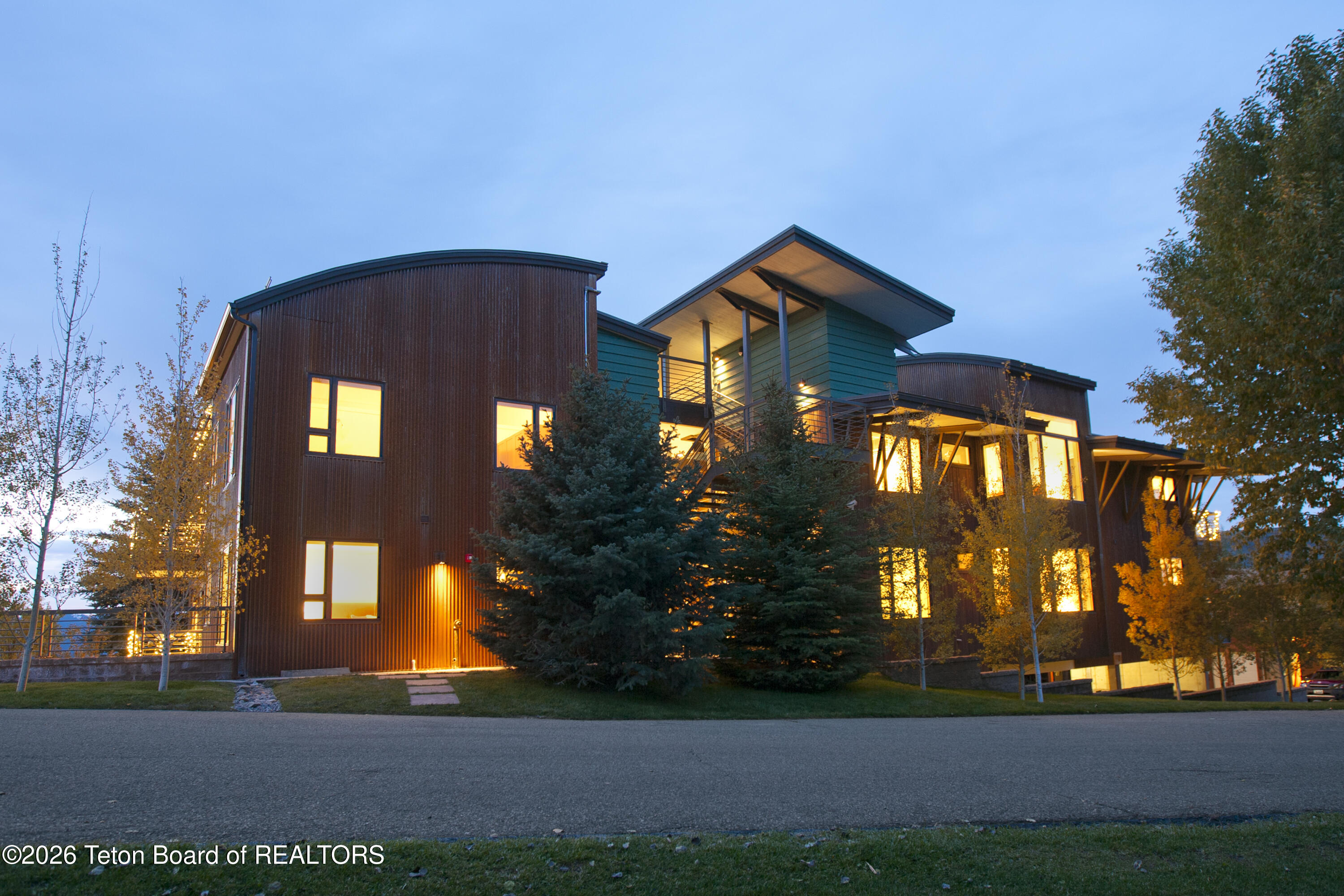 Modern multi-story home with wood siding, covered balcony, and warmly lit windows at dusk.