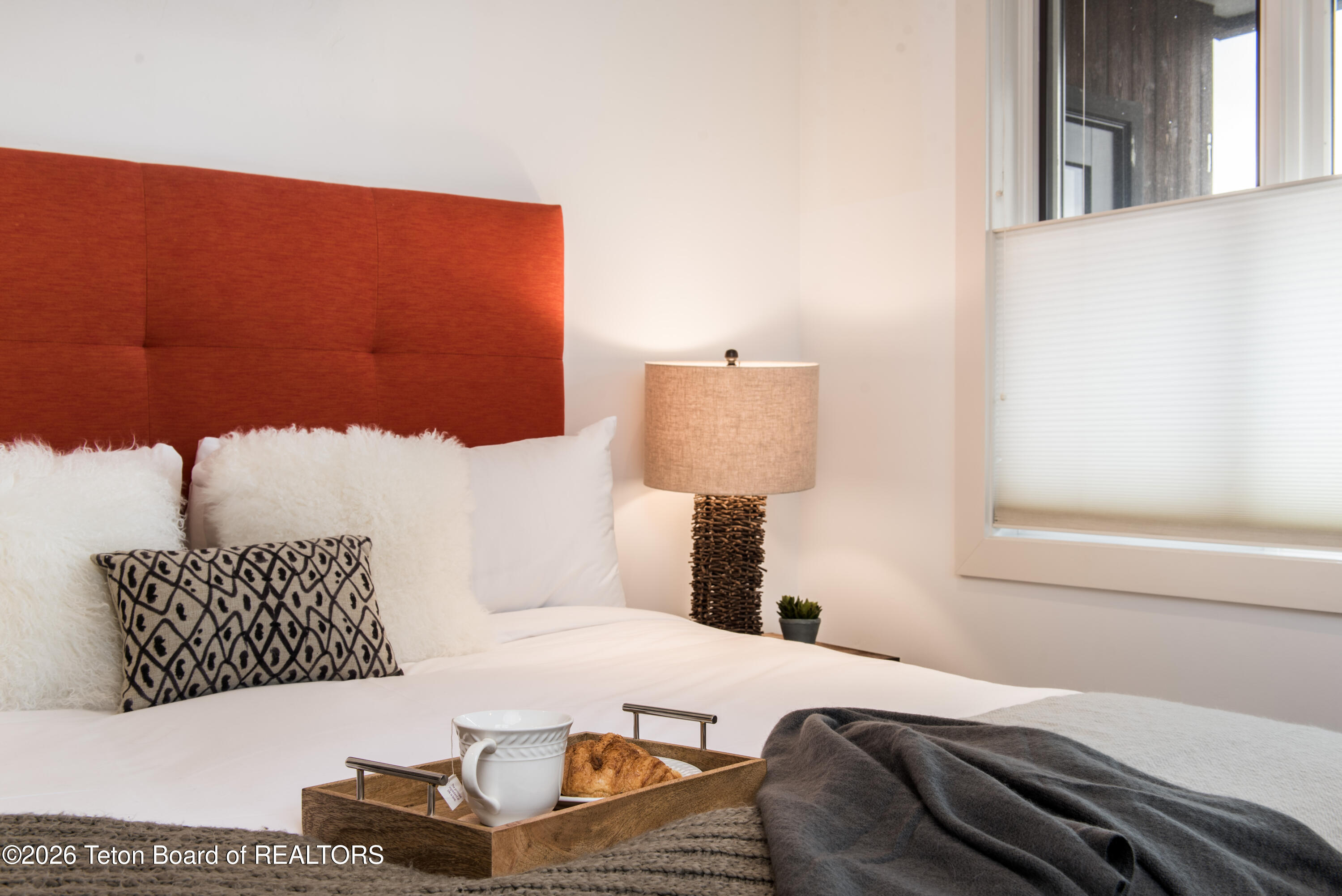 Staged bedroom with a rust-colored headboard, layered white pillows, and a breakfast tray on the bed.