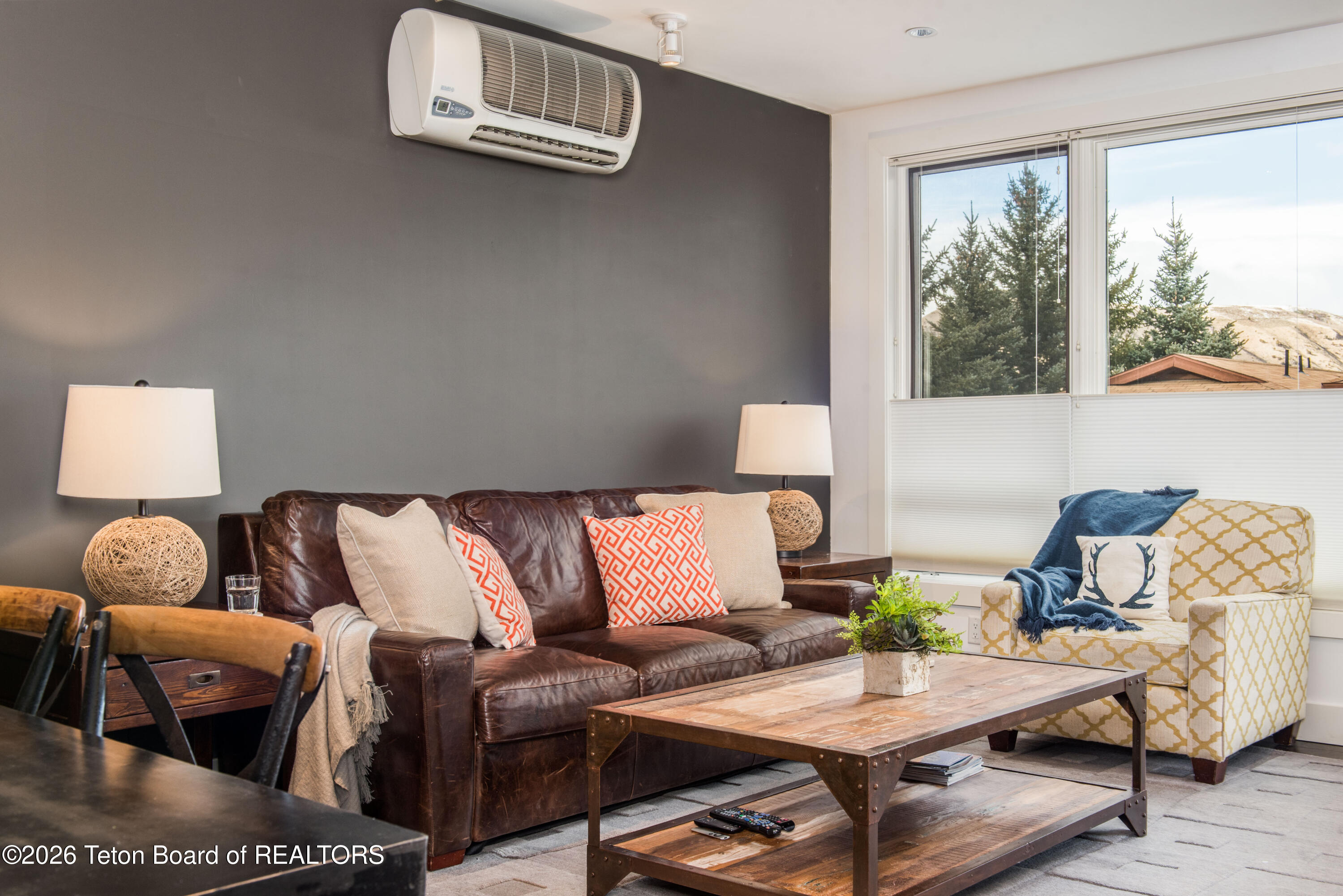 Living room with a leather sofa, patterned armchair, rustic coffee table, and window overlooking evergreen trees.