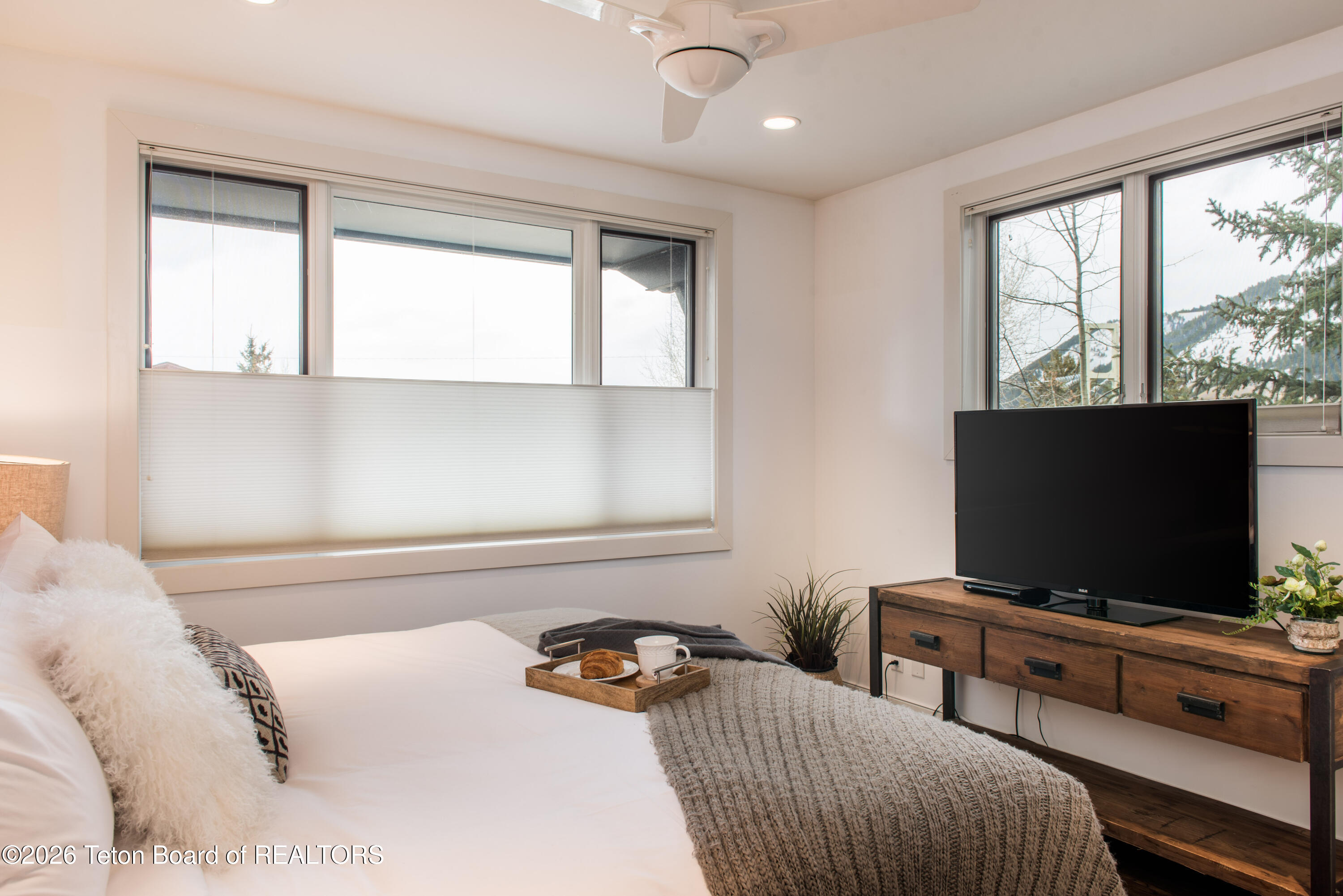 Bedroom with a white bed, gray throw, television, and snowy mountain views through large windows.