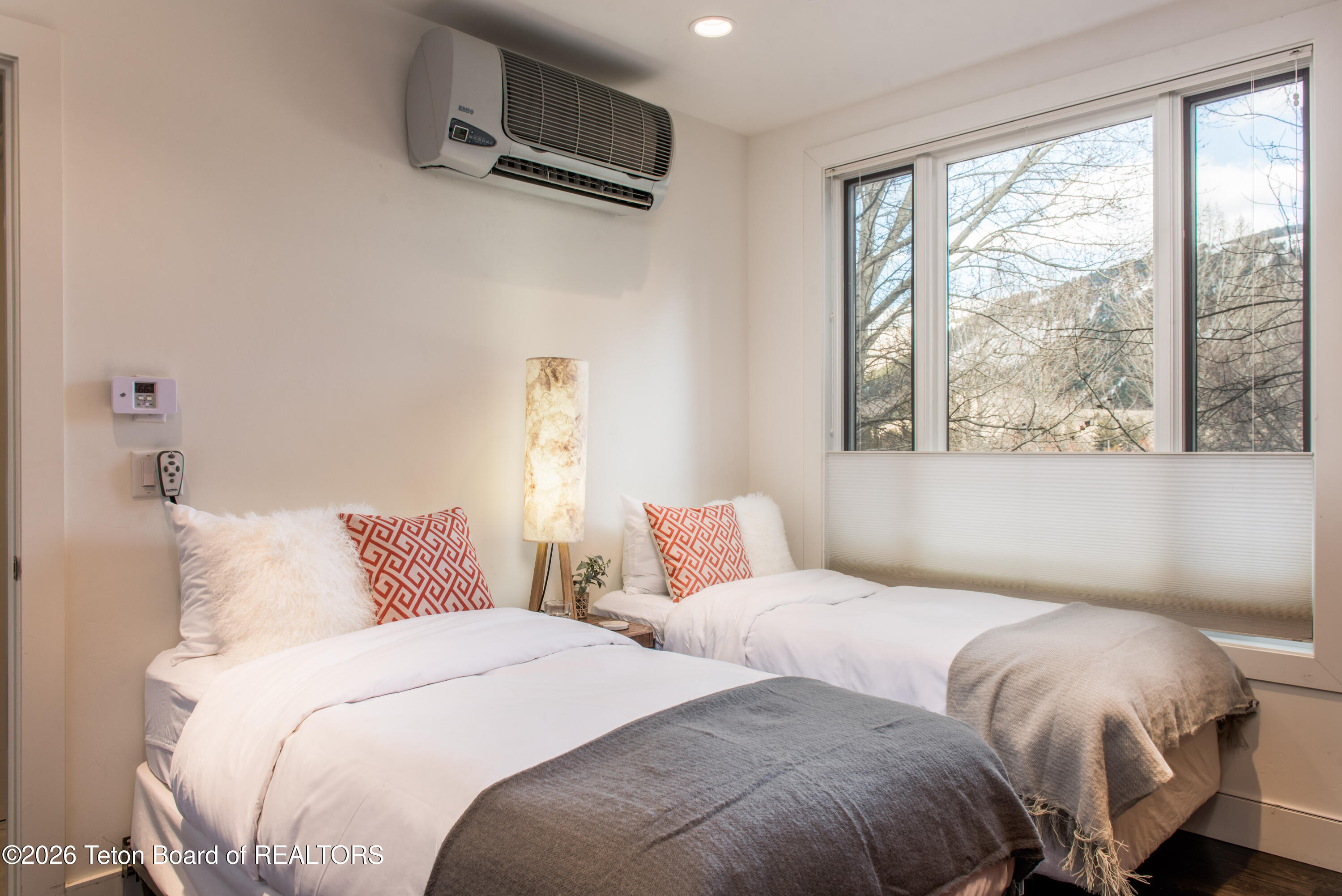 Bedroom with two separate beds, orange patterned pillows, and a large window overlooking leafless trees.