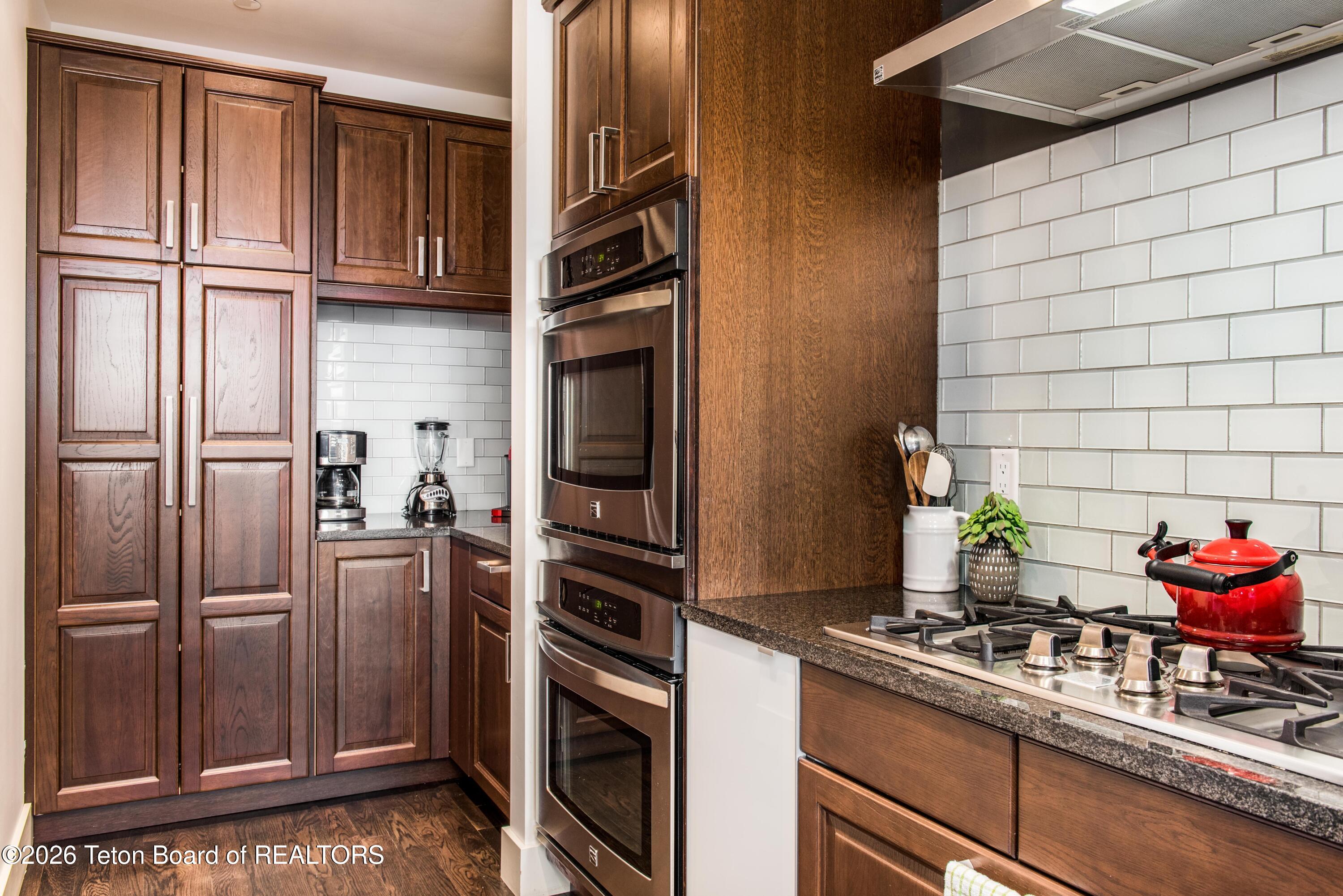 Kitchen with dark wood cabinetry, double wall ovens, and a gas cooktop beneath a stainless steel hood.