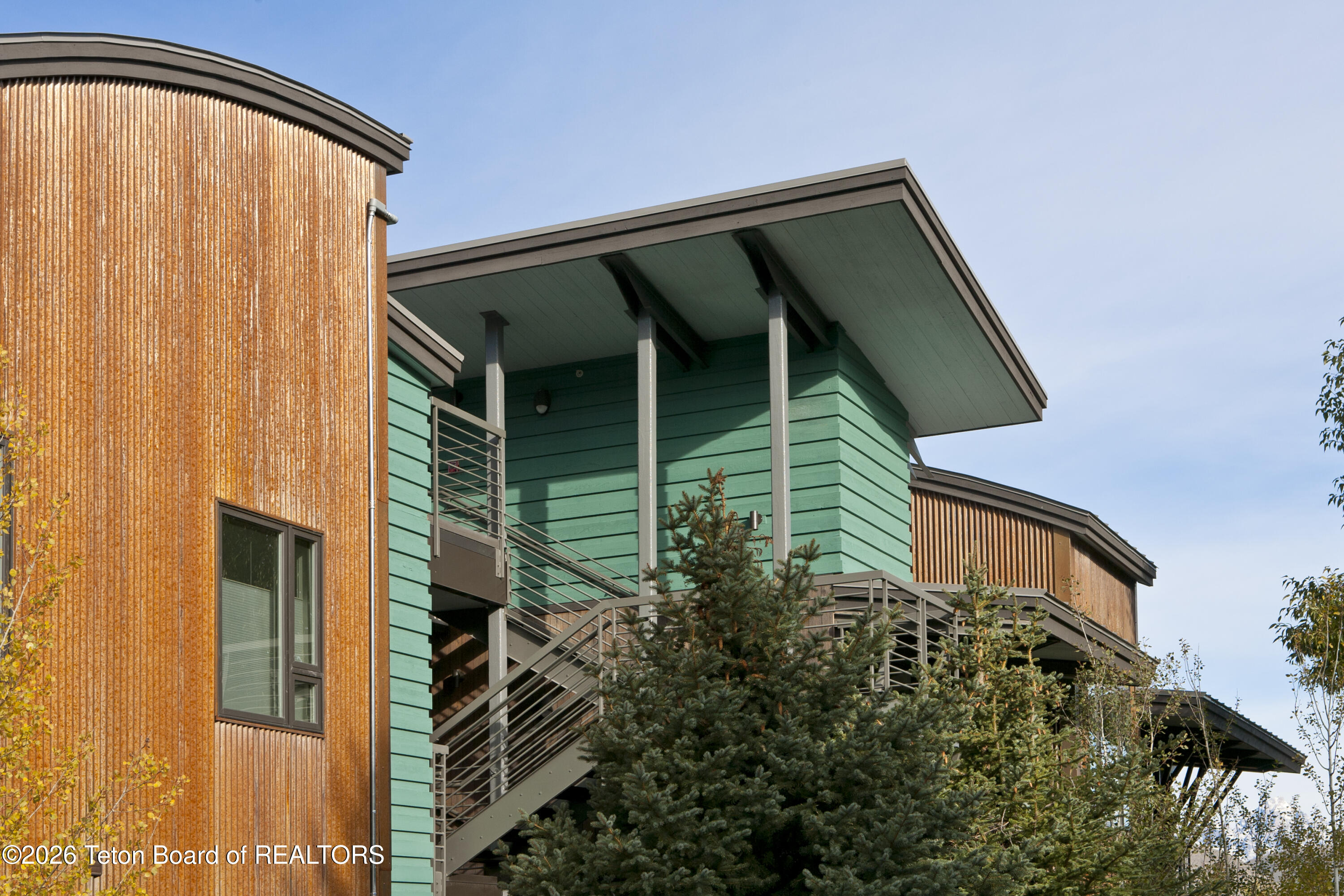 Modern residence with teal siding, wood cladding, and an exterior staircase among evergreen trees.