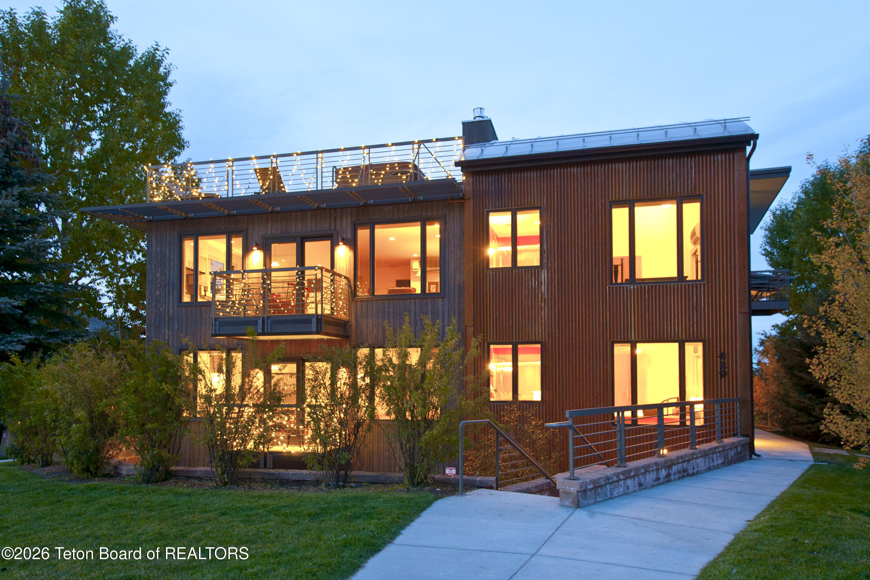 Modern wood-clad home at dusk with a rooftop terrace outlined in lights and a projecting balcony.