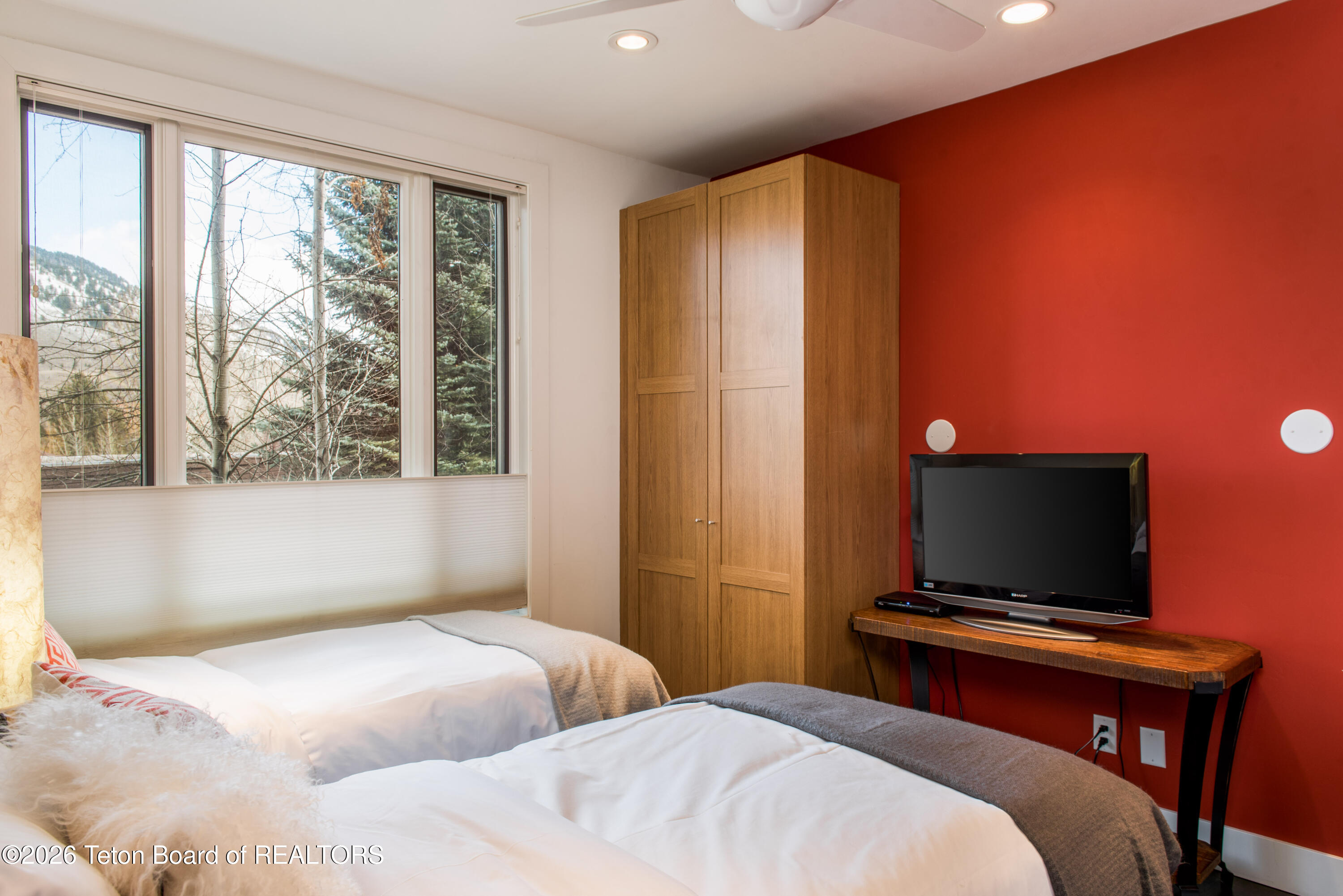 Bedroom with two beds, a wooden wardrobe, and a red accent wall, with mountain views through the window.