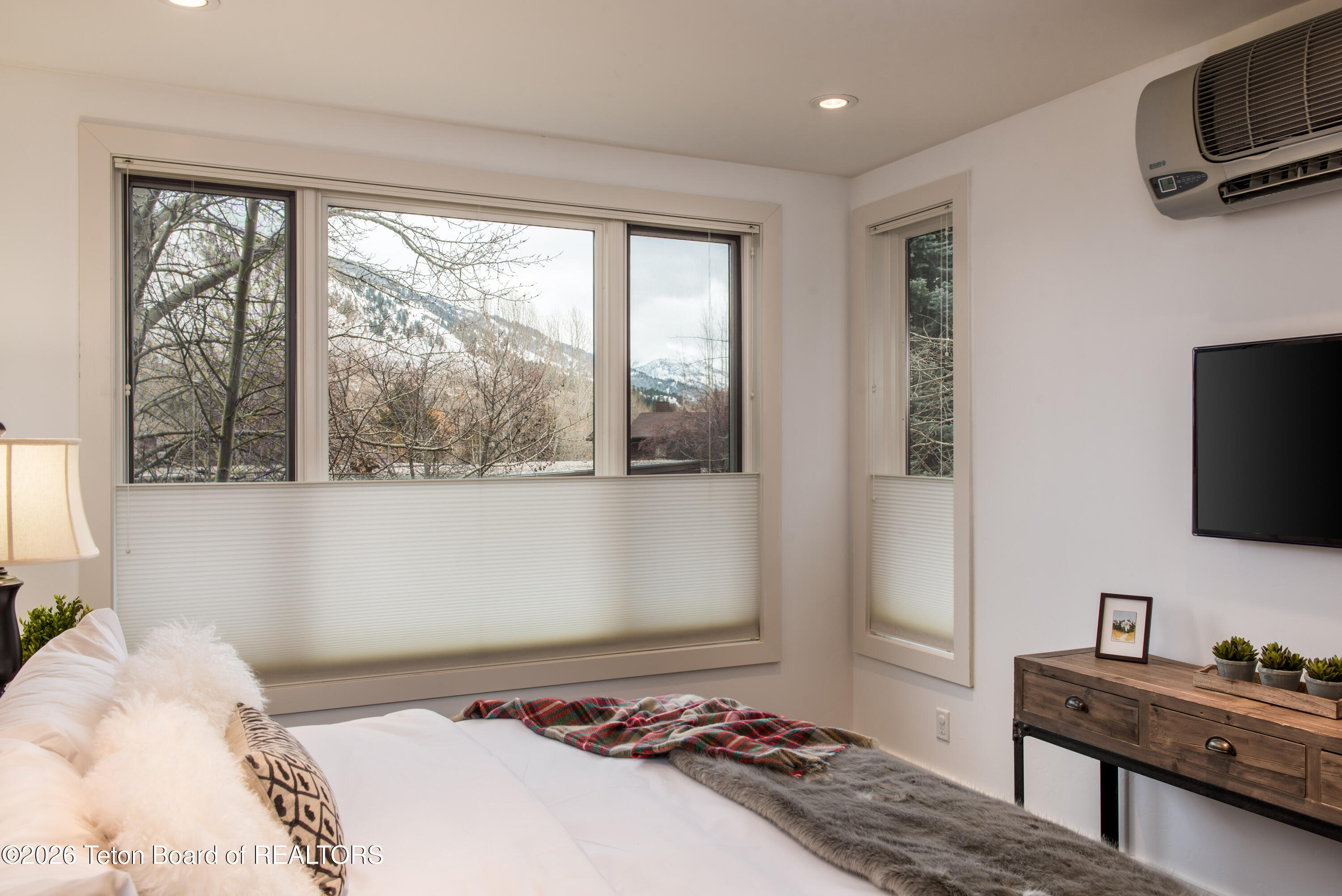 Furnished bedroom with wide windows overlooking snow-covered mountains, a plaid blanket, and a wooden dresser.
