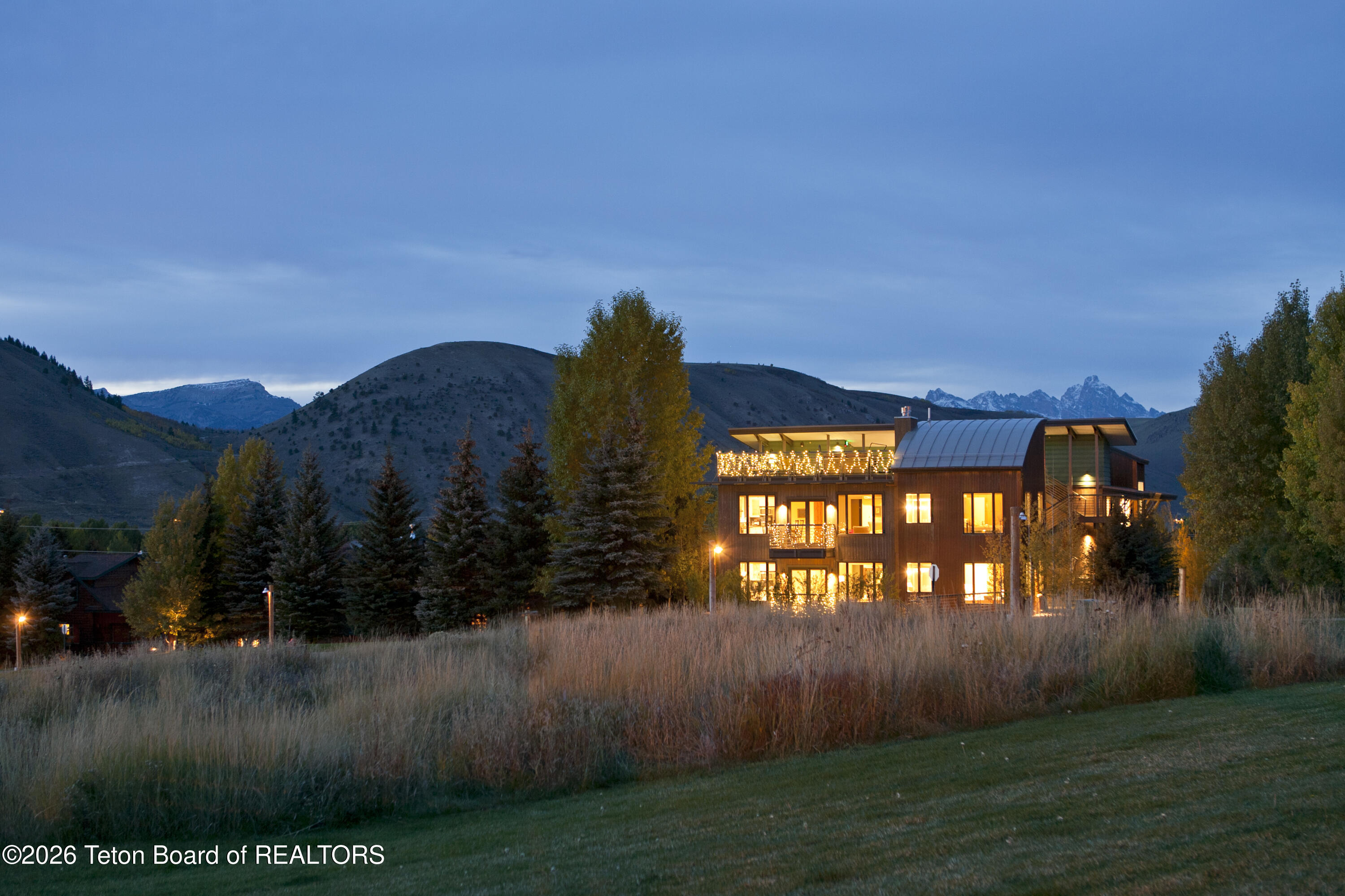 Modern mountain home glowing at dusk, with a lit rooftop terrace and autumn grasses.