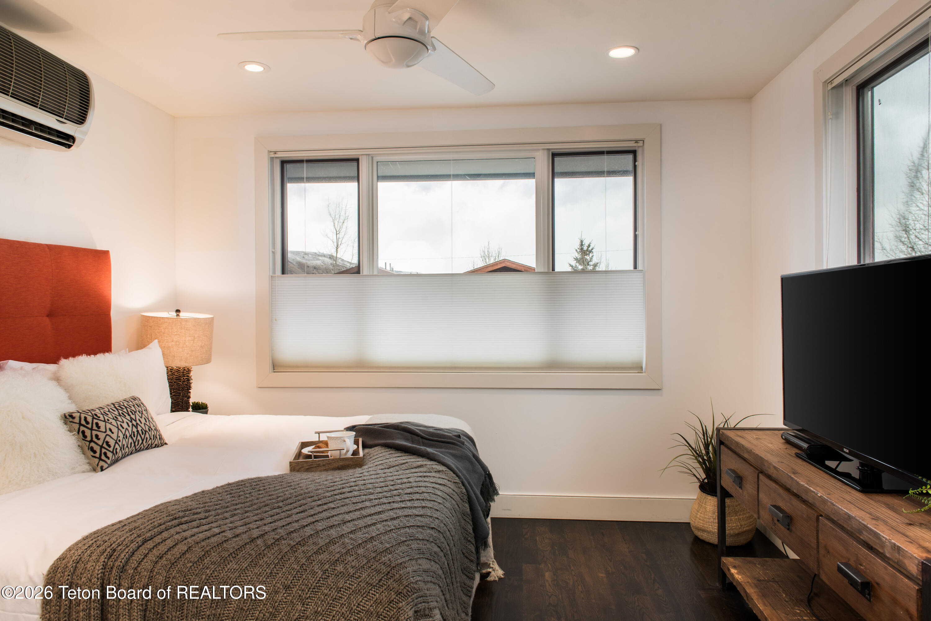 Staged bedroom with a rust-colored headboard, gray knit blanket, and large shaded window.