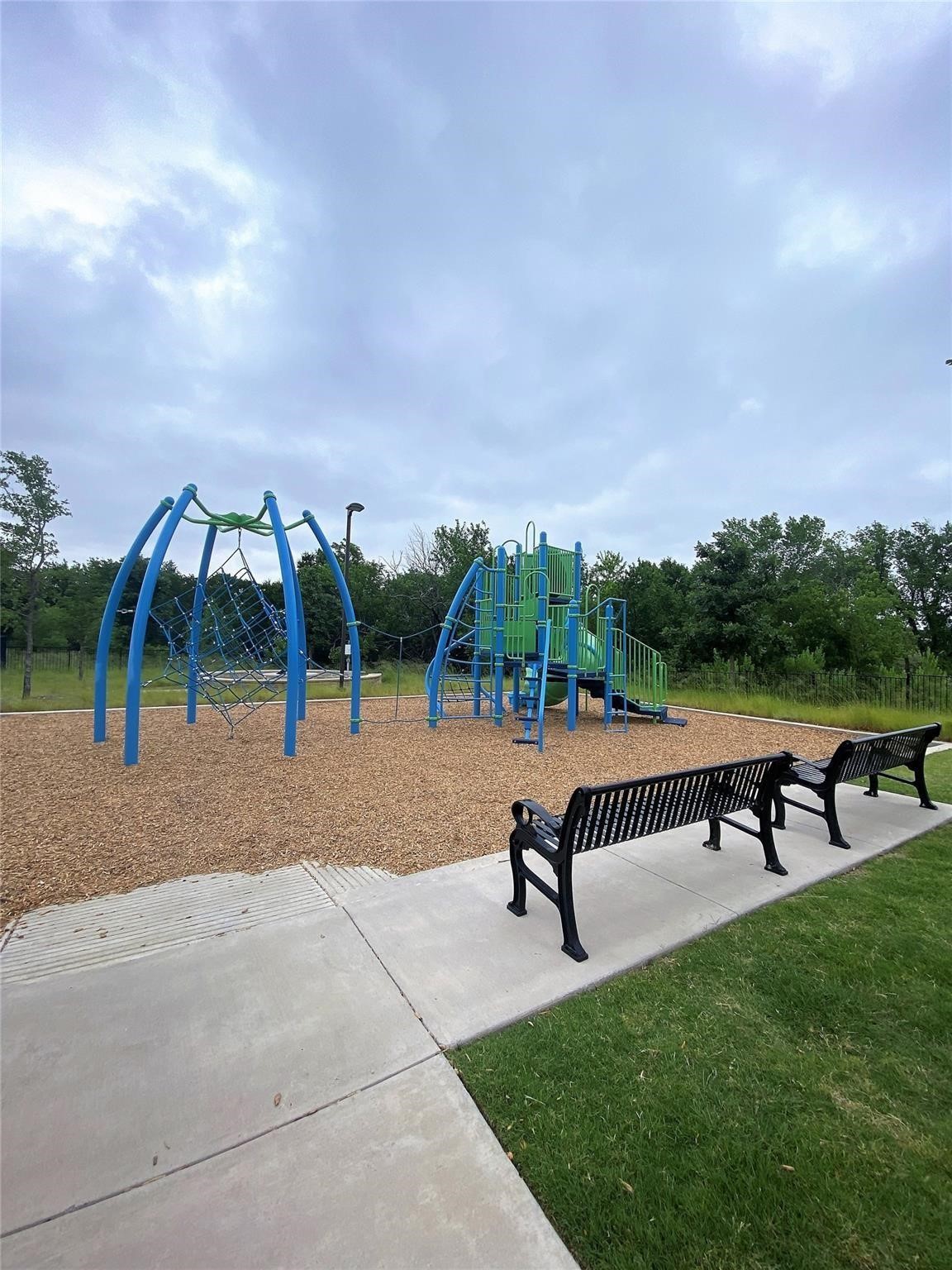 Community playground with blue and green climbing equipment and benches along a paved path.
