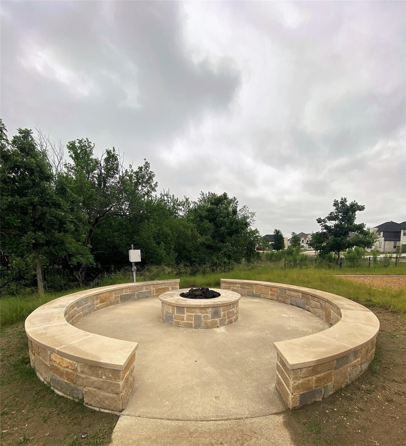 Circular stone fire pit seating area beside trees and a fenced lawn.