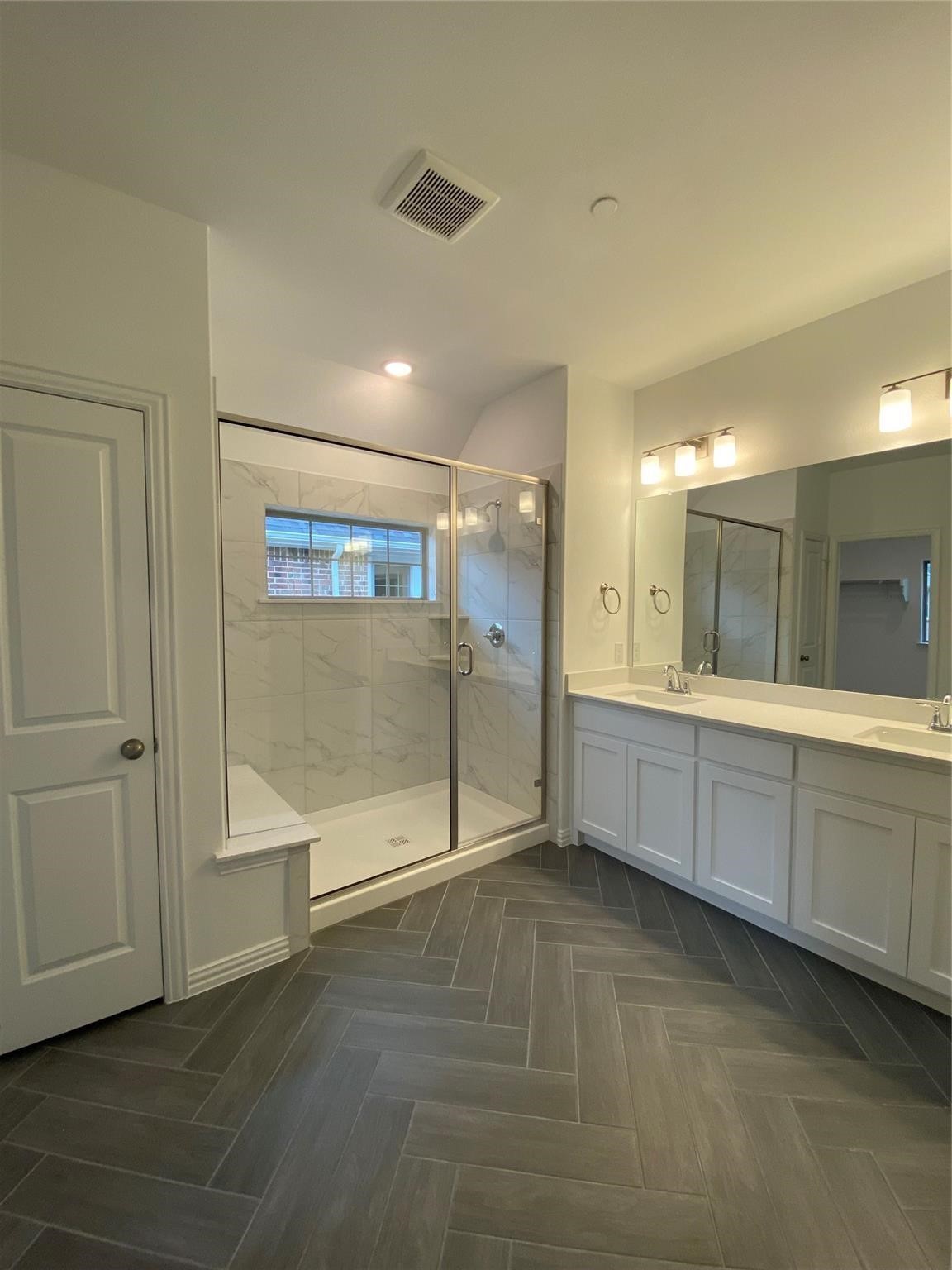 Bathroom with a glass-enclosed tiled shower, long double vanity, and dark gray herringbone floor.