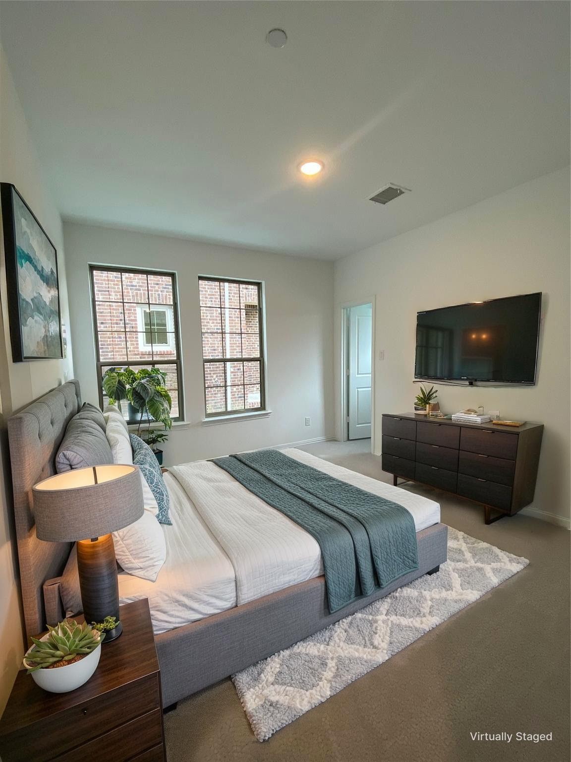Staged bedroom with a gray upholstered bed, teal bedding, two tall windows, and a wall-mounted television.
