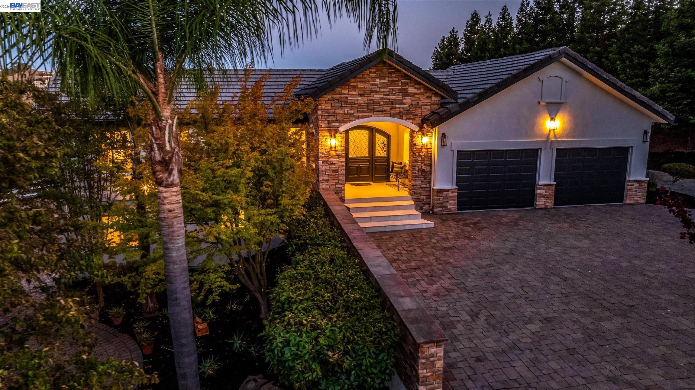 Stone-and-stucco home at dusk with a warmly lit entry, double garage, and palm-lined landscaping.