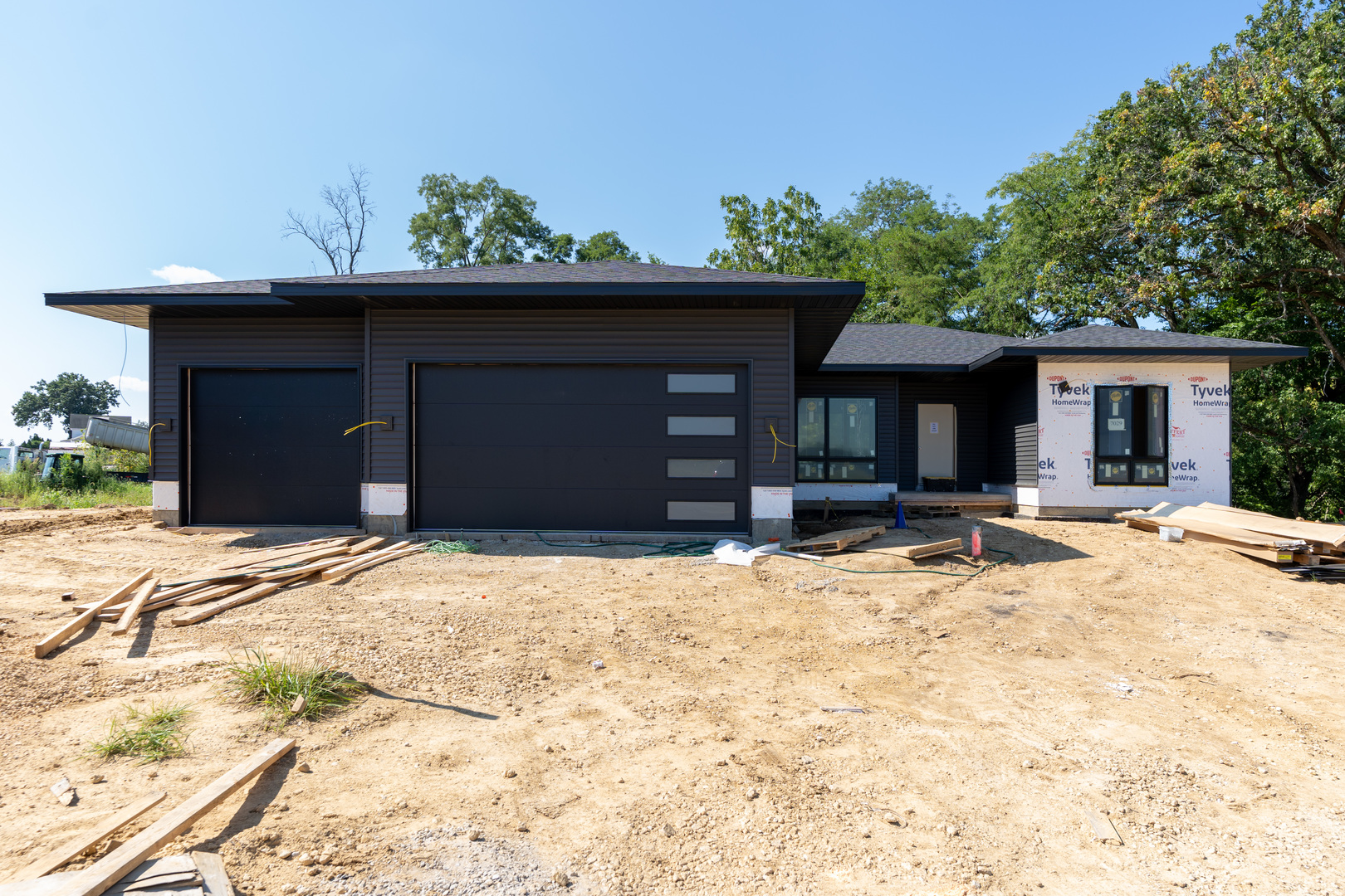 Modern single-story home under construction with dark siding, broad garage doors, and unfinished ground.