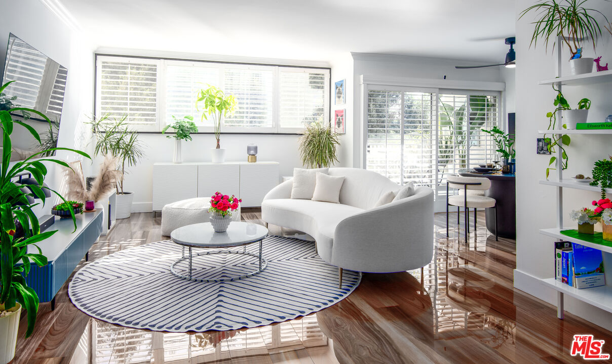 Bright living room with a curved white sofa, patterned rug, glossy floors, and large shuttered windows.