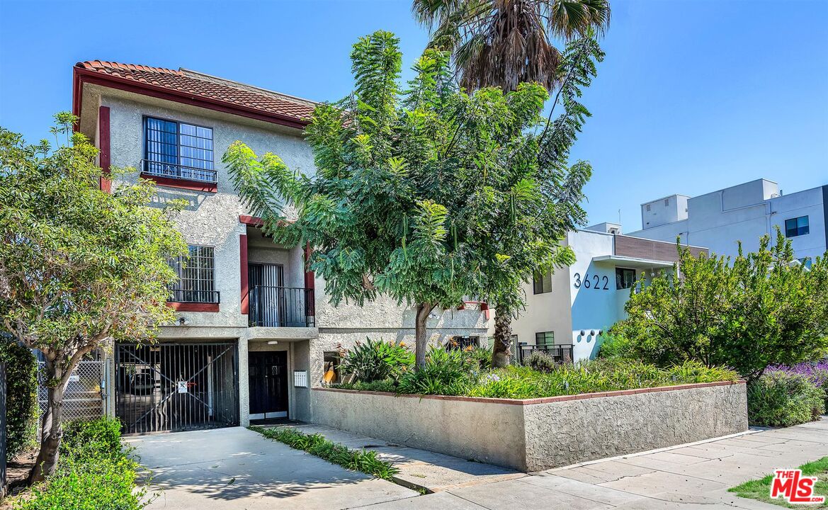 Two-story stucco apartment building with red trim, gated carport, and mature trees along the sidewalk.