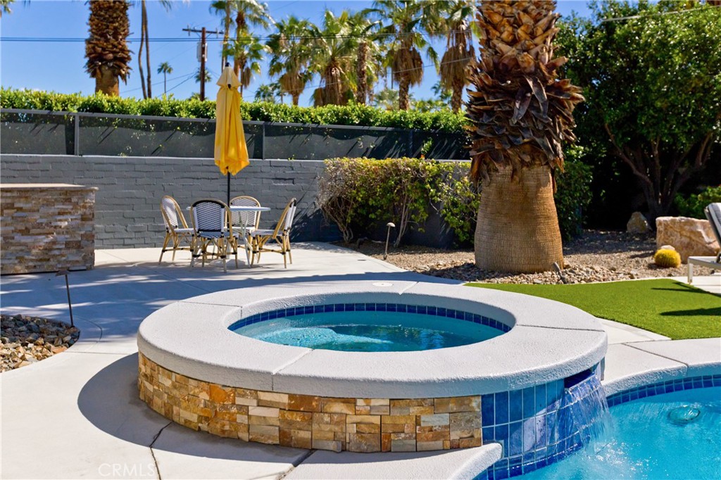 Raised circular spa spills into a pool beside a patio dining set and palm trees.