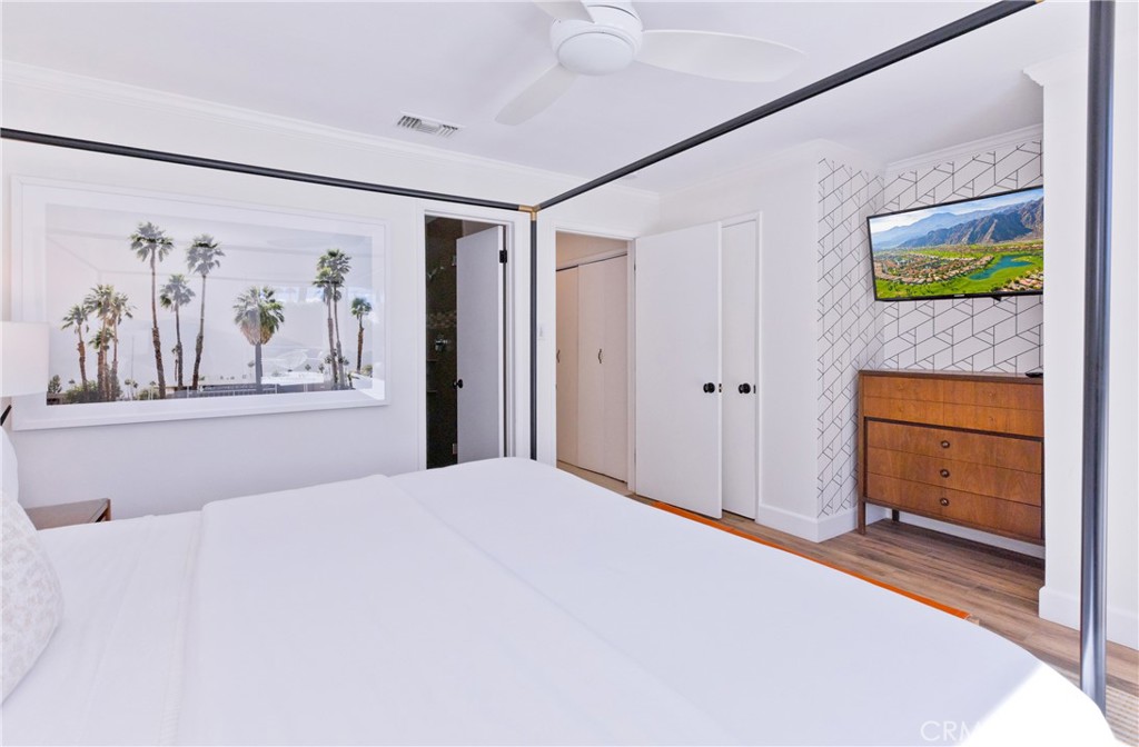 Bedroom with a large white bed, framed palm-tree photograph, and wooden dresser beneath a television.