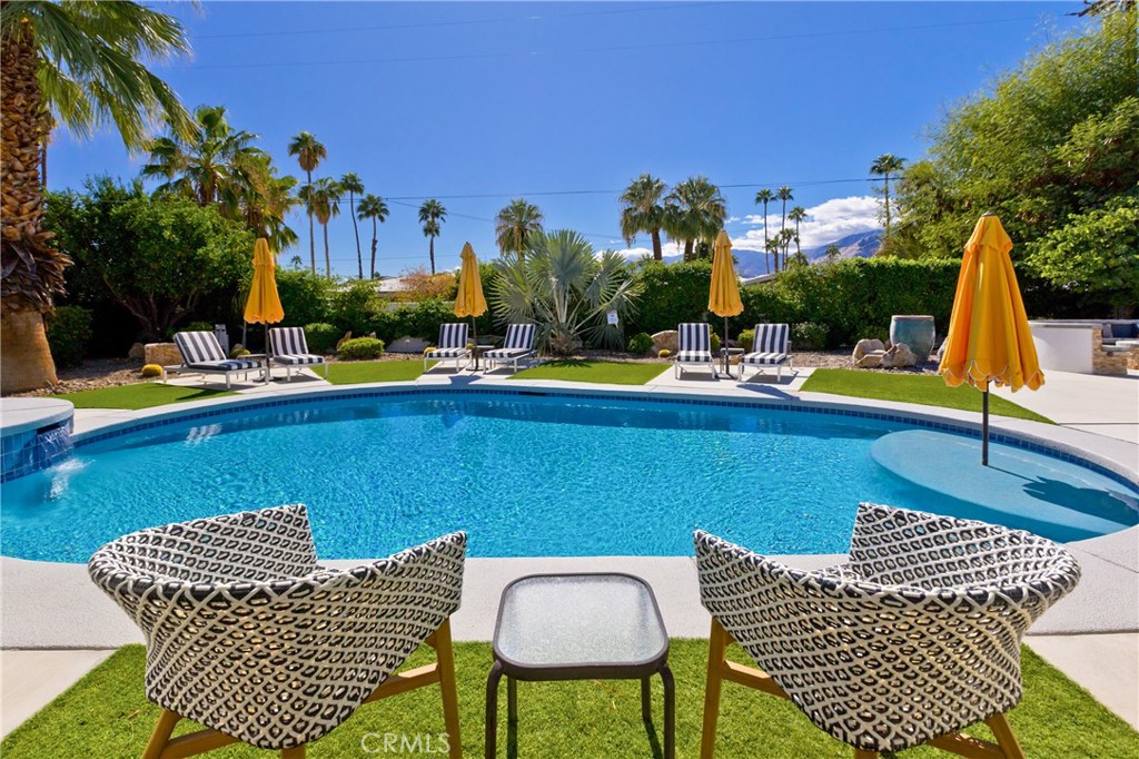 Backyard pool with patterned lounge chairs, yellow umbrellas, palm trees, and distant mountains.