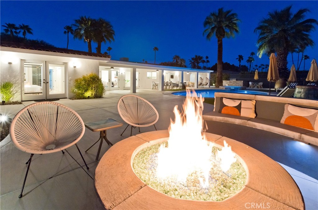 Modern home patio at night with a pool, circular fire pit, and palm trees.