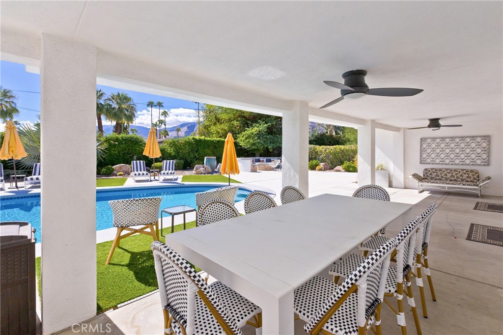 Covered patio dining area overlooking a pool, lawn, palm trees, and distant mountains.