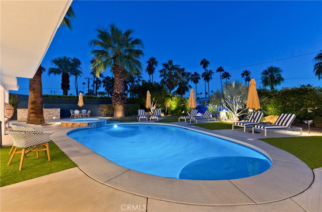 Backyard swimming pool surrounded by striped lounge chairs, palm trees, and illuminated landscaping at dusk.