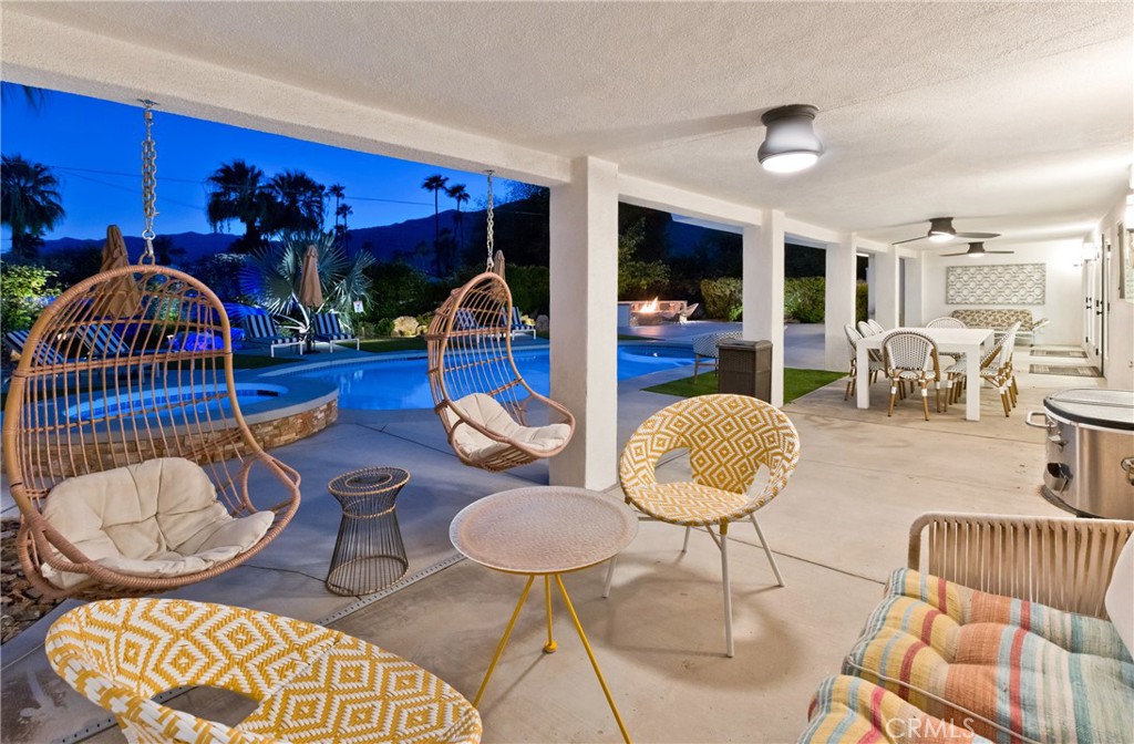 Covered patio with hanging wicker chairs and dining area beside a lit swimming pool.