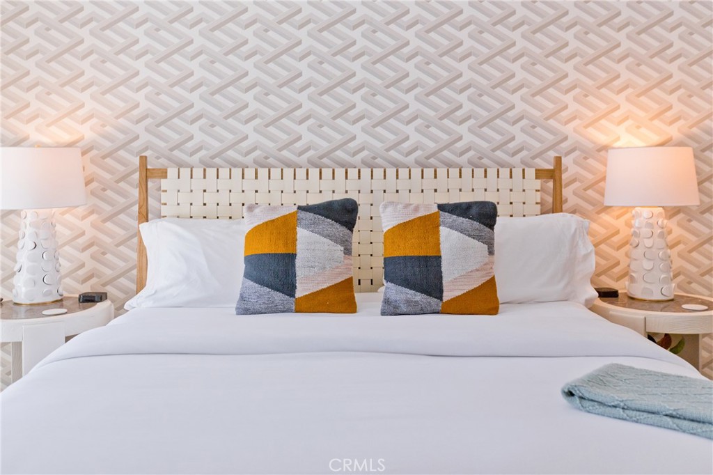 Staged bedroom with a white bed, geometric accent pillows, woven headboard, and patterned wallpaper.
