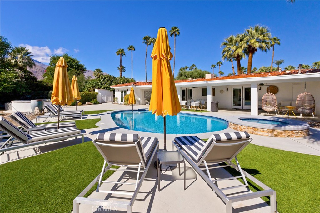 Backyard pool and raised spa with striped loungers and yellow umbrellas beside a palm-framed home.
