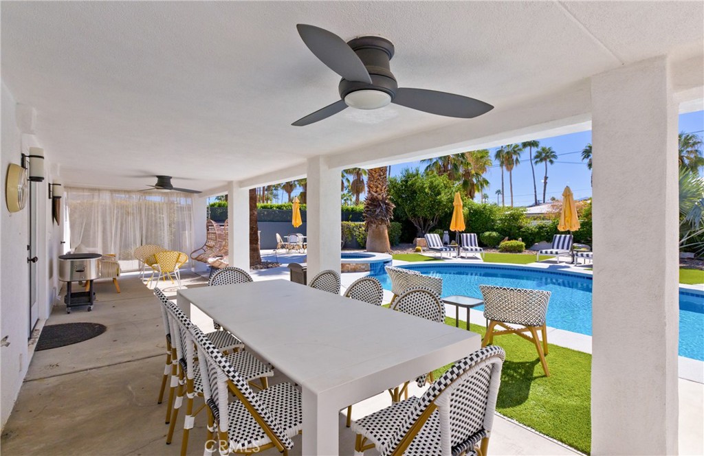 Covered patio with a long dining table beside a pool, palm trees, and yellow umbrellas.