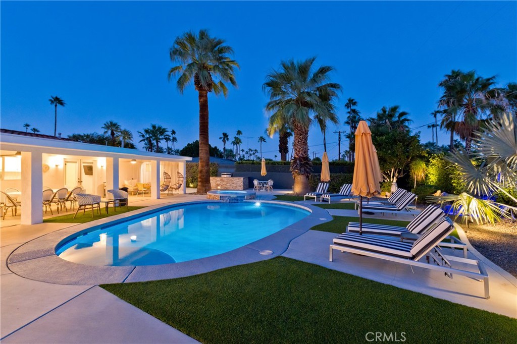 Backyard pool at dusk with palm trees, striped loungers, and a brightly lit covered patio.