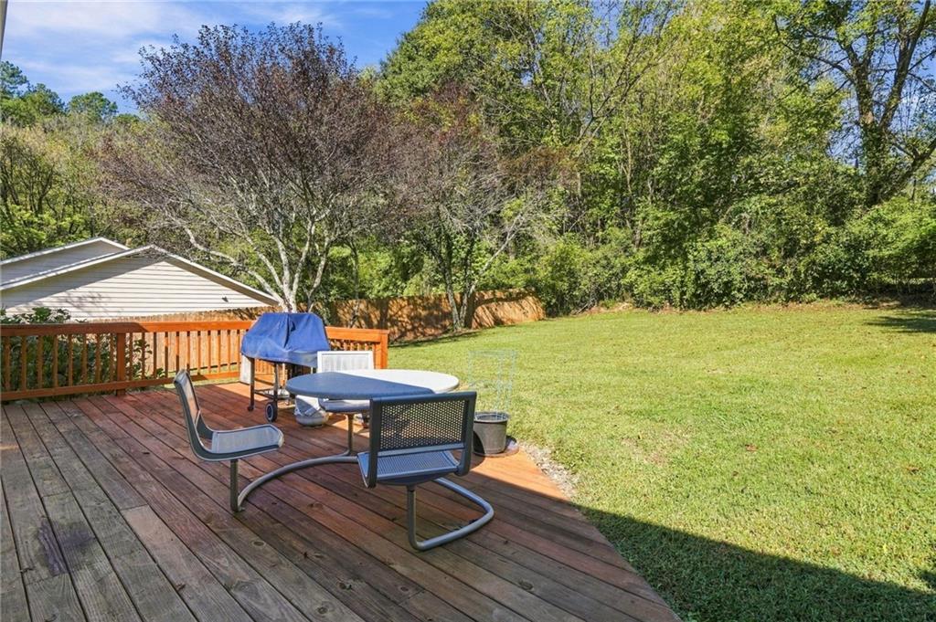 Wooden deck with outdoor dining furniture and a grill overlooks a sunny lawn bordered by trees.