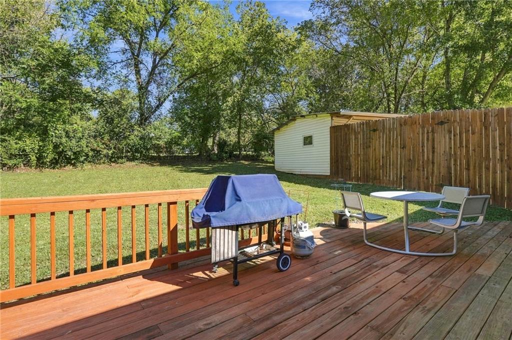 Wooden backyard deck with a covered grill, patio chairs, and a fenced lawn beneath mature trees.