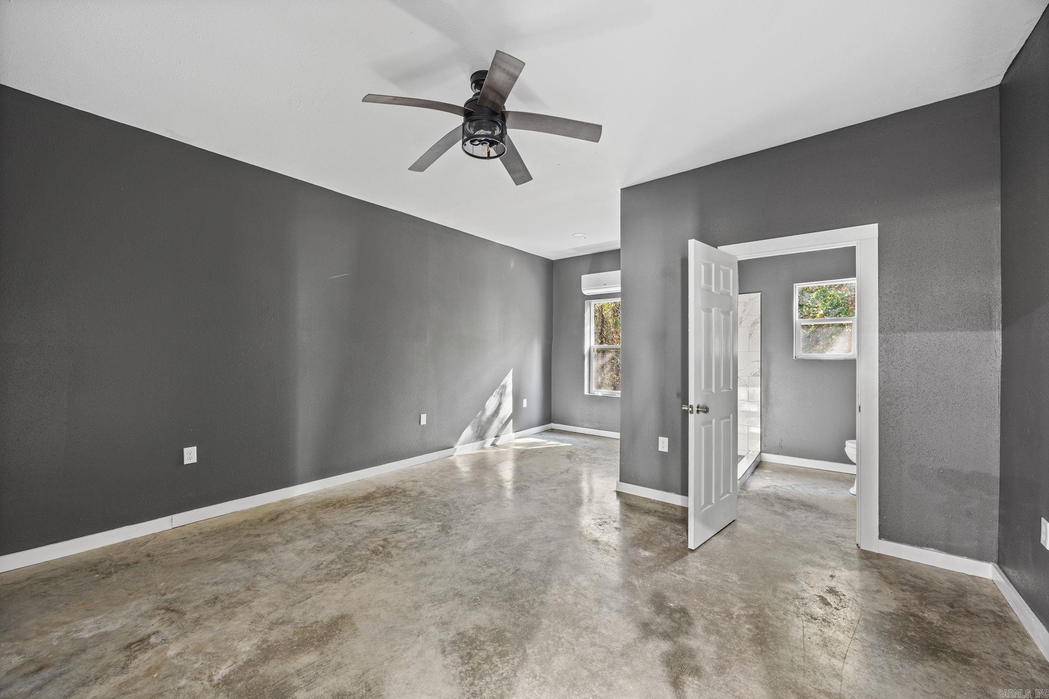 Empty gray room with a concrete floor, ceiling fan, and open doorway to a bathroom.