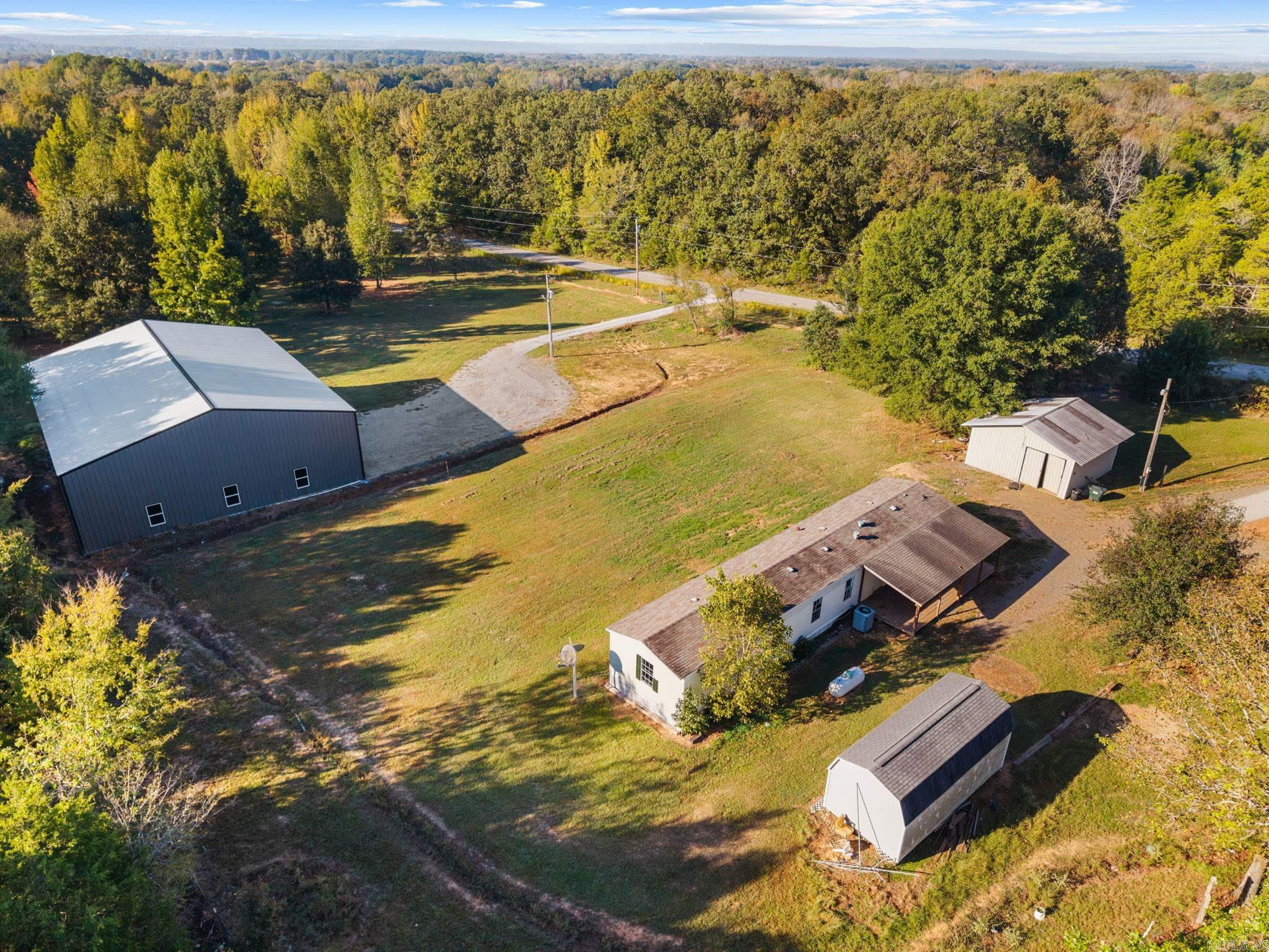 Aerial view of a rural property with a home, multiple outbuildings, open lawn, and surrounding woods.