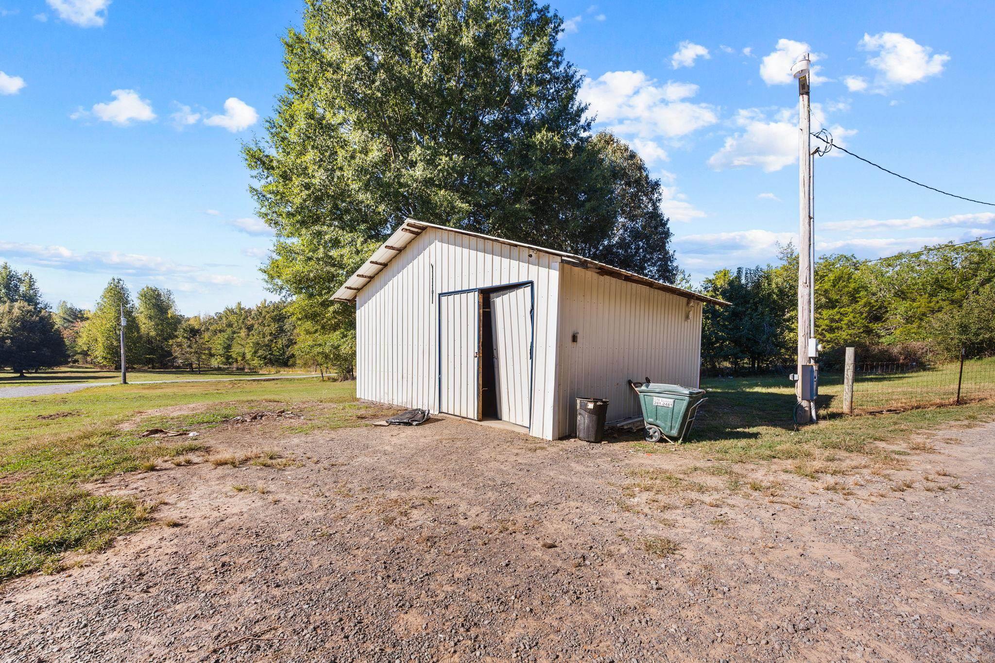 White metal outbuilding with an open doorway beside a gravel yard and mature trees.