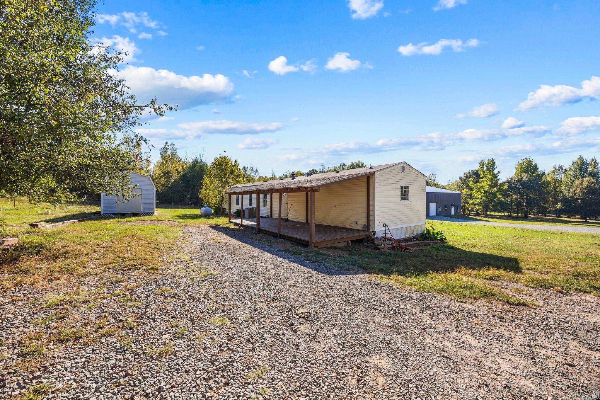 Single-story home with a long covered porch, gravel driveway, and detached outbuildings.