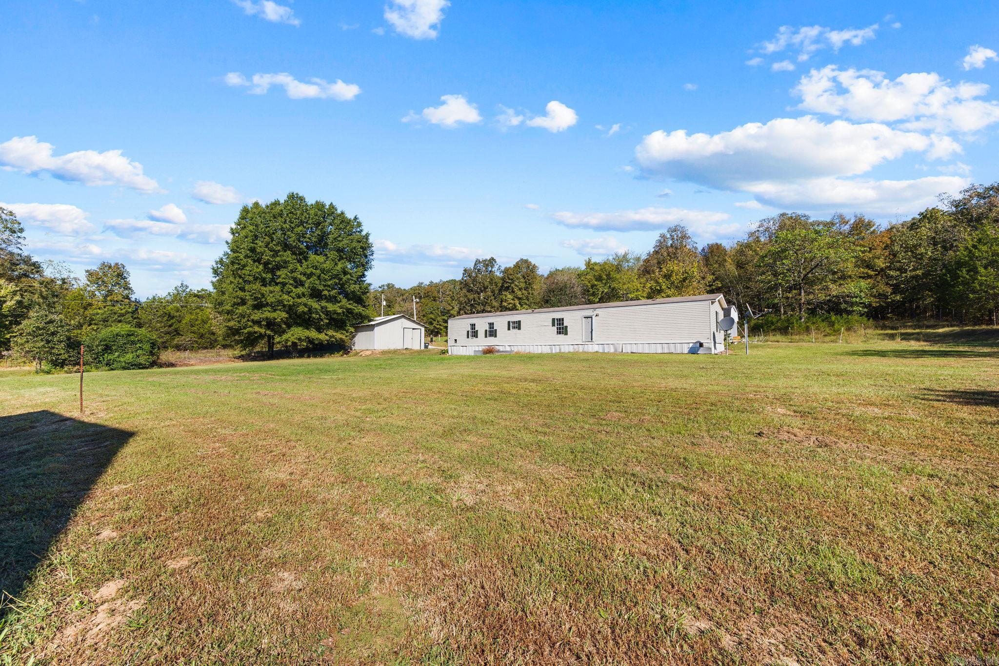 Long manufactured home and small outbuilding beyond a broad grassy yard and mature trees.