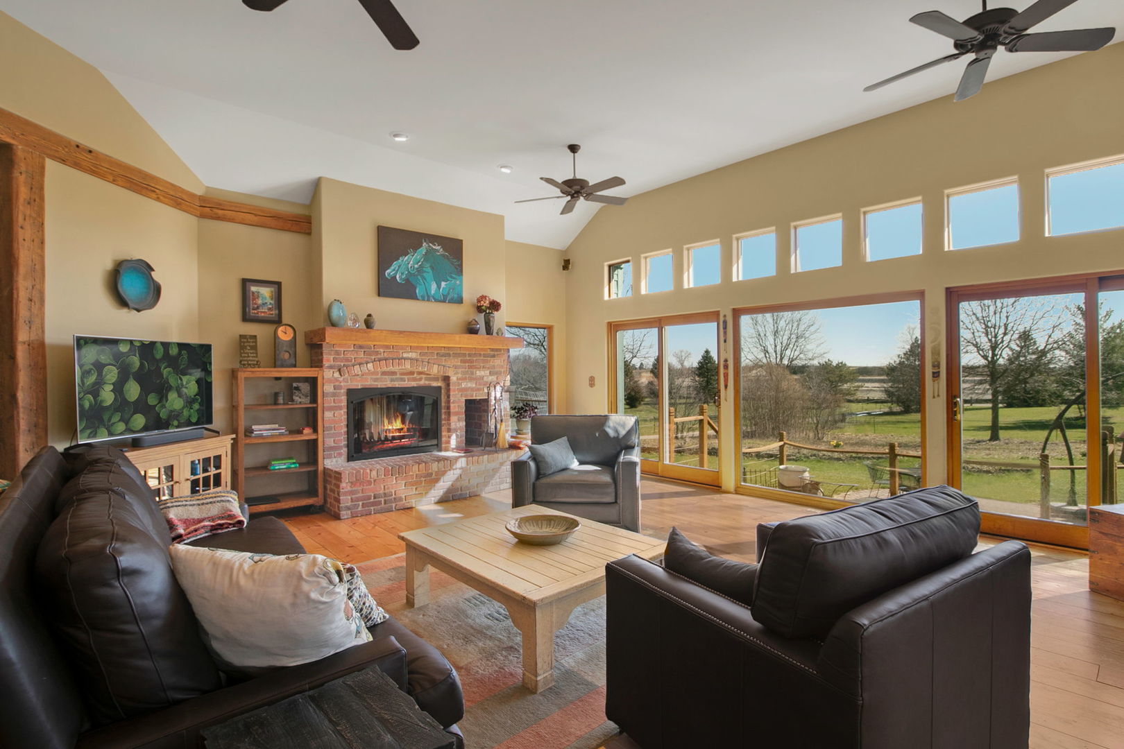 Living room with a brick fireplace, vaulted ceiling, and large windows overlooking green grounds.