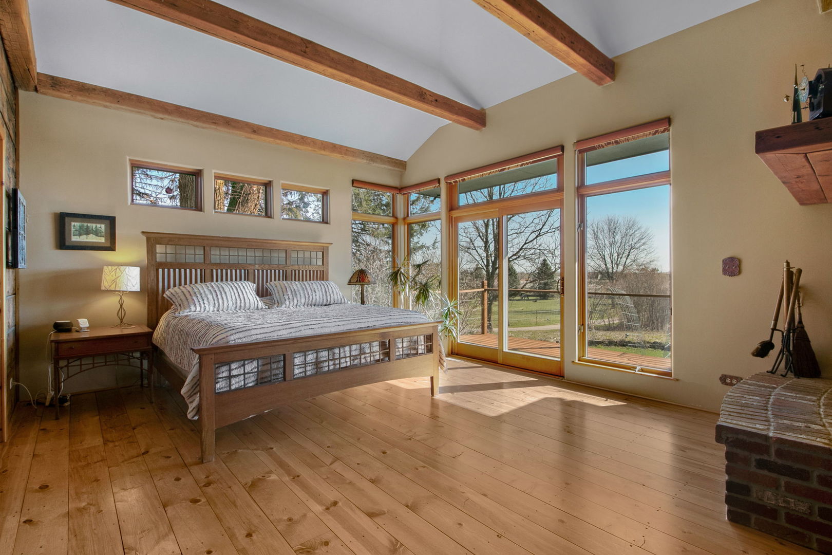 Bedroom with exposed wood beams, plank floors, and tall windows opening onto a deck.