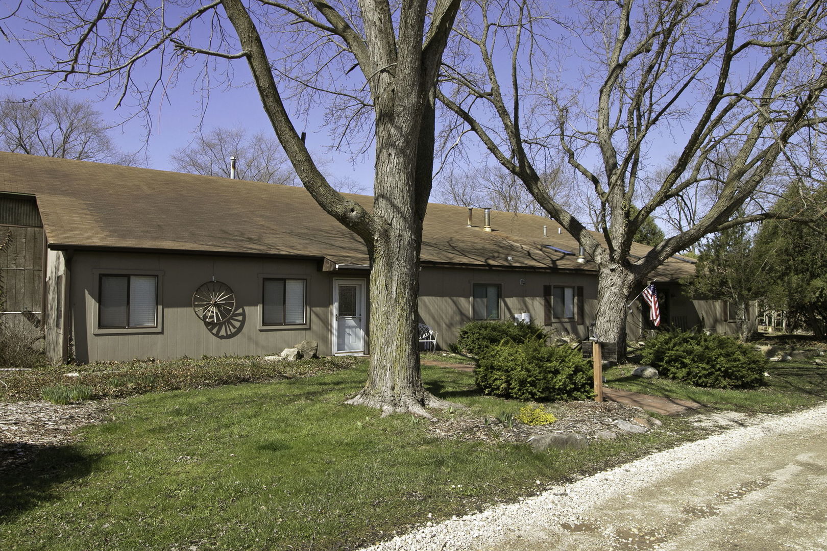 Single-story brown home framed by large bare trees, with a gravel drive and American flag.