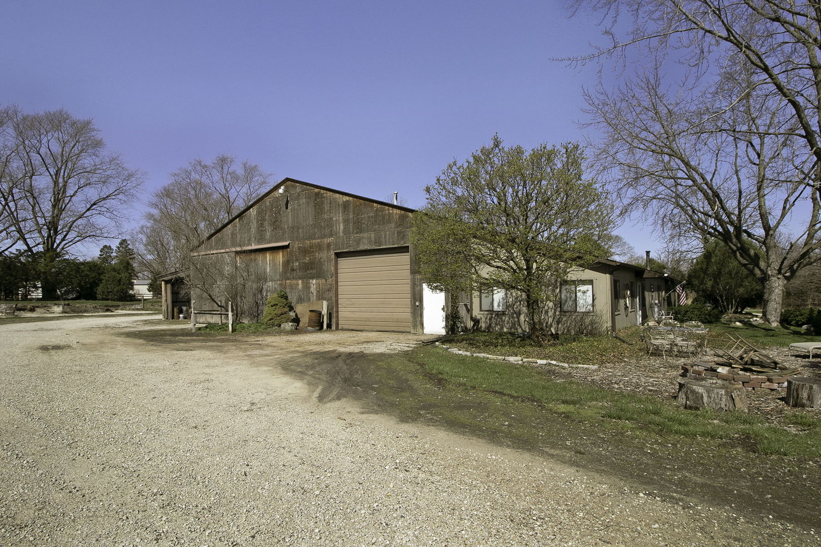 One-story home beside a large weathered barn, with a gravel driveway and mature trees.