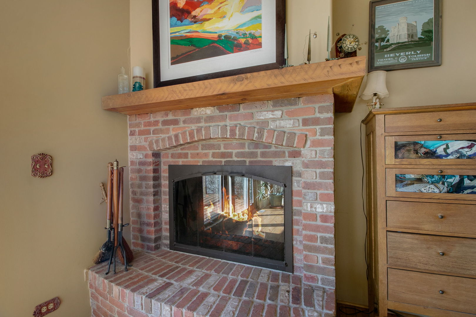 Brick fireplace with a glass-front firebox, broad wooden mantel, and fireplace tools beside the hearth.