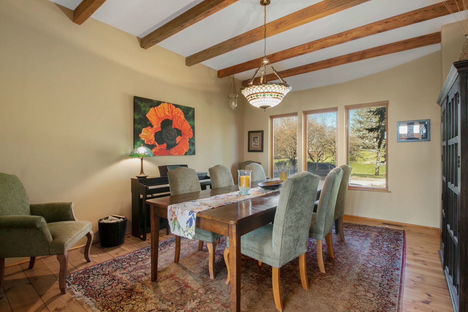 Dining room with exposed wooden beams, a long table, and tall windows overlooking the yard.