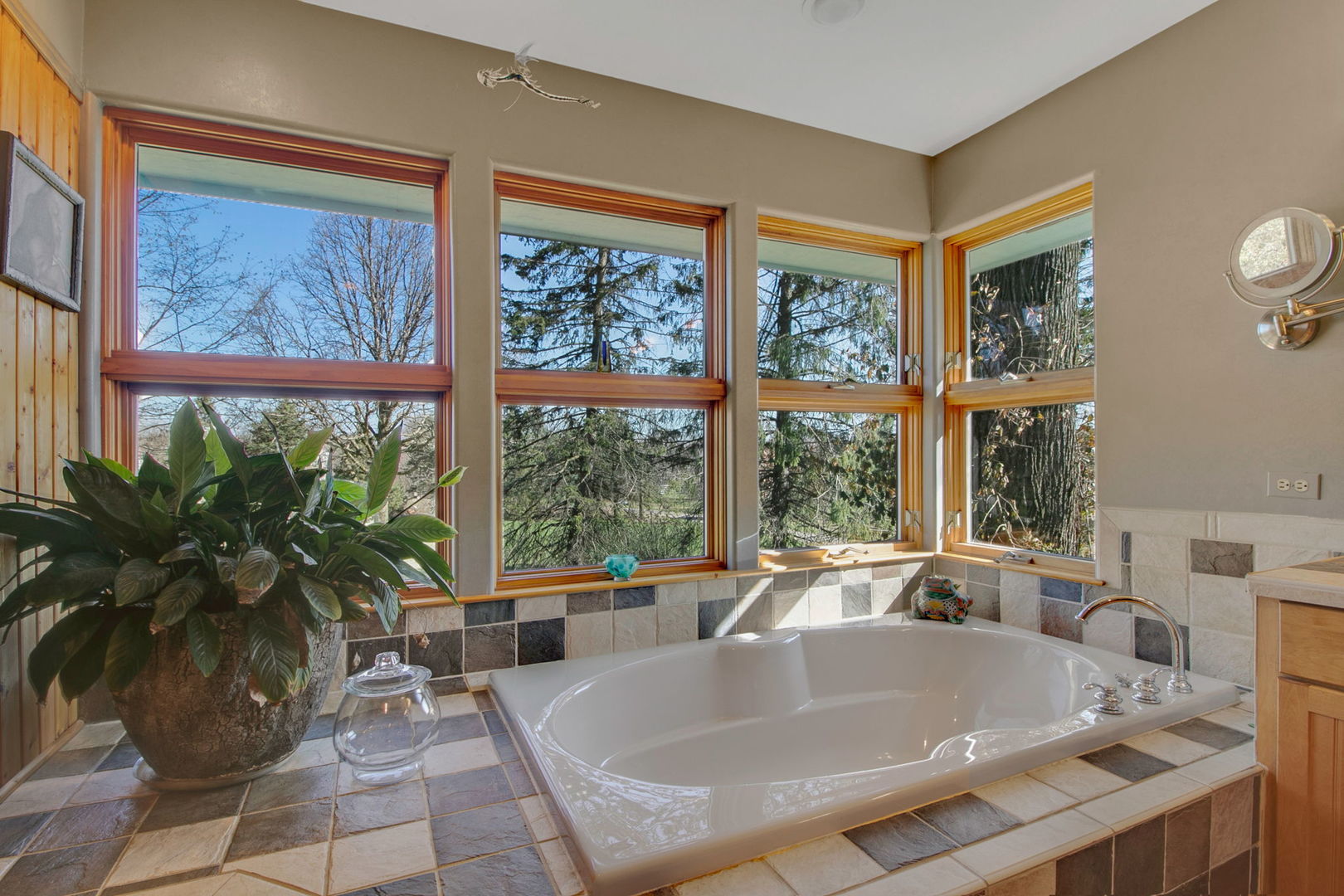 Bathroom soaking tub beneath tall windows, with tiled surround and trees visible outside.