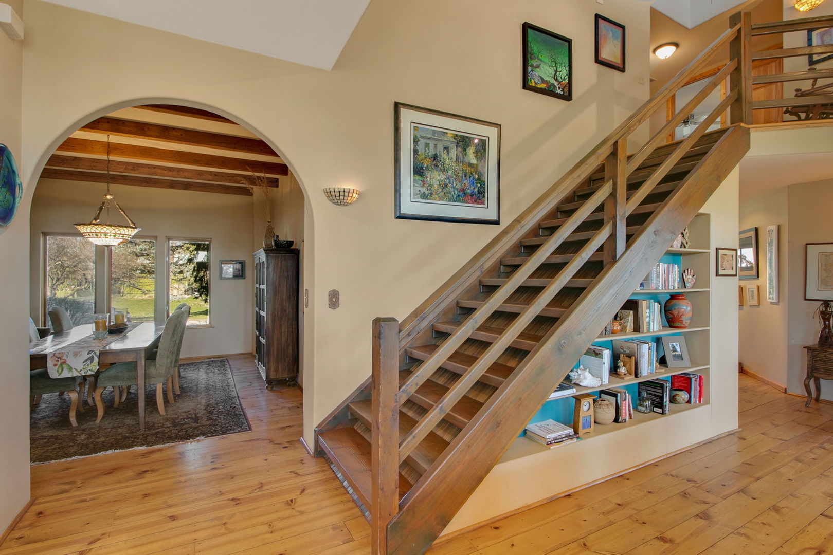 Wooden staircase with built-in bookshelves beside an arched dining area with exposed beams.