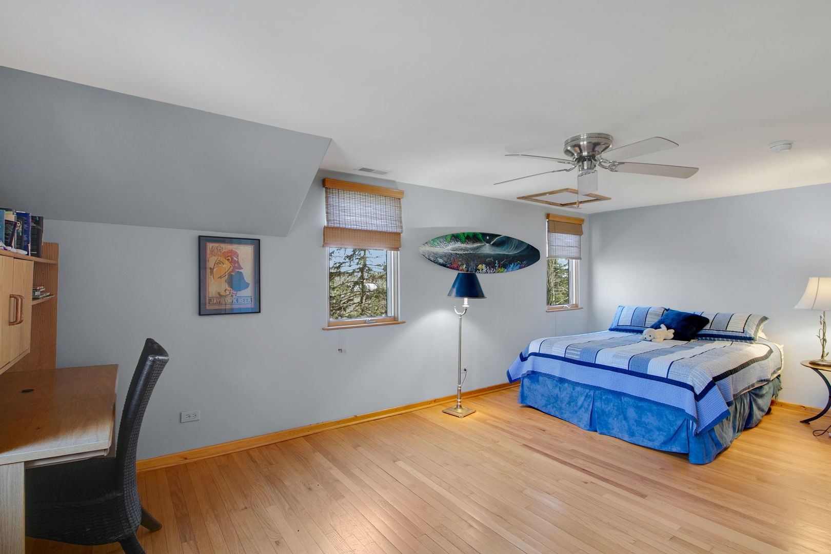 Bedroom with a blue-and-white bedspread, hardwood floors, and two windows with woven shades.