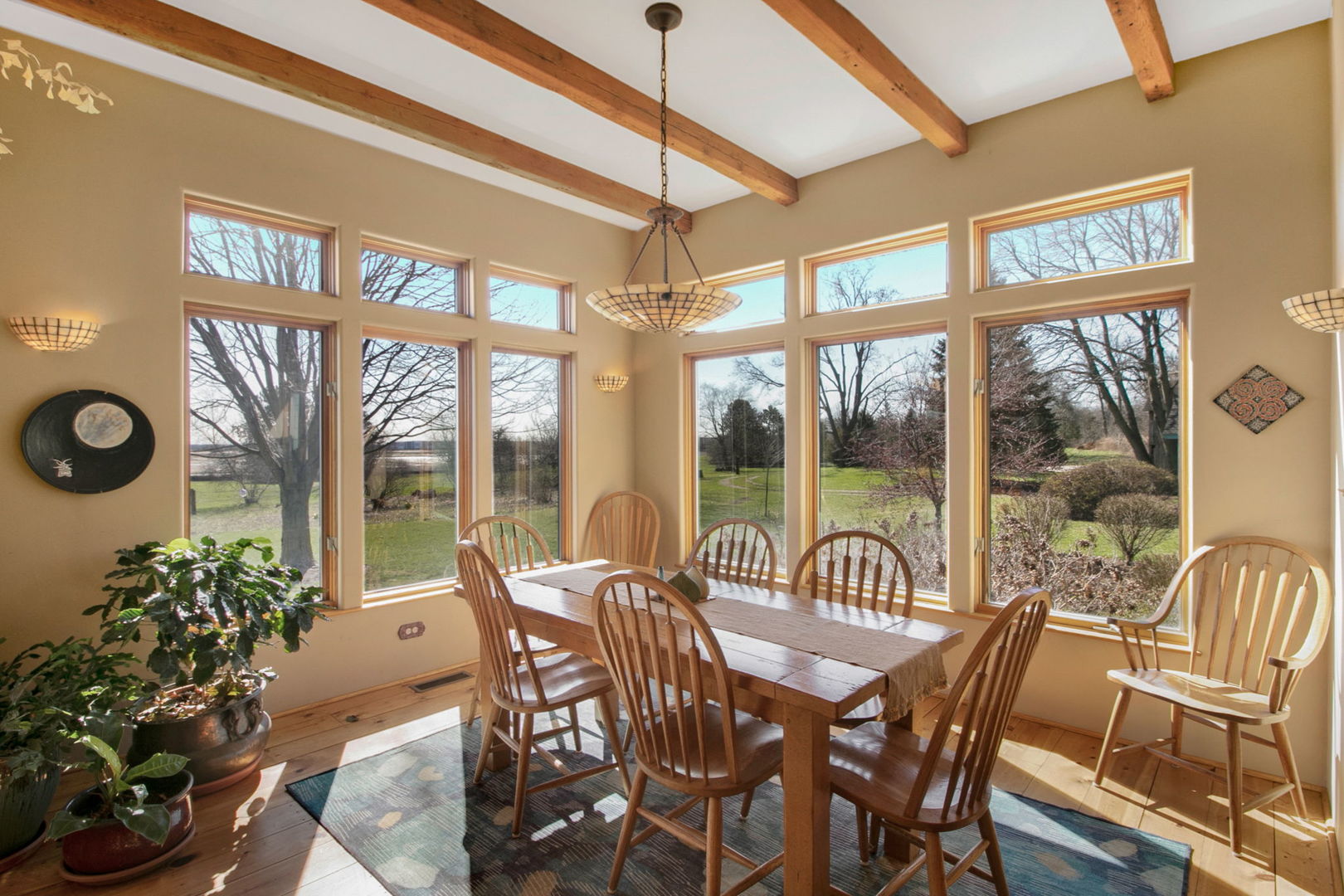Dining room with exposed timber beams, a long wooden table, and tall windows overlooking the yard.