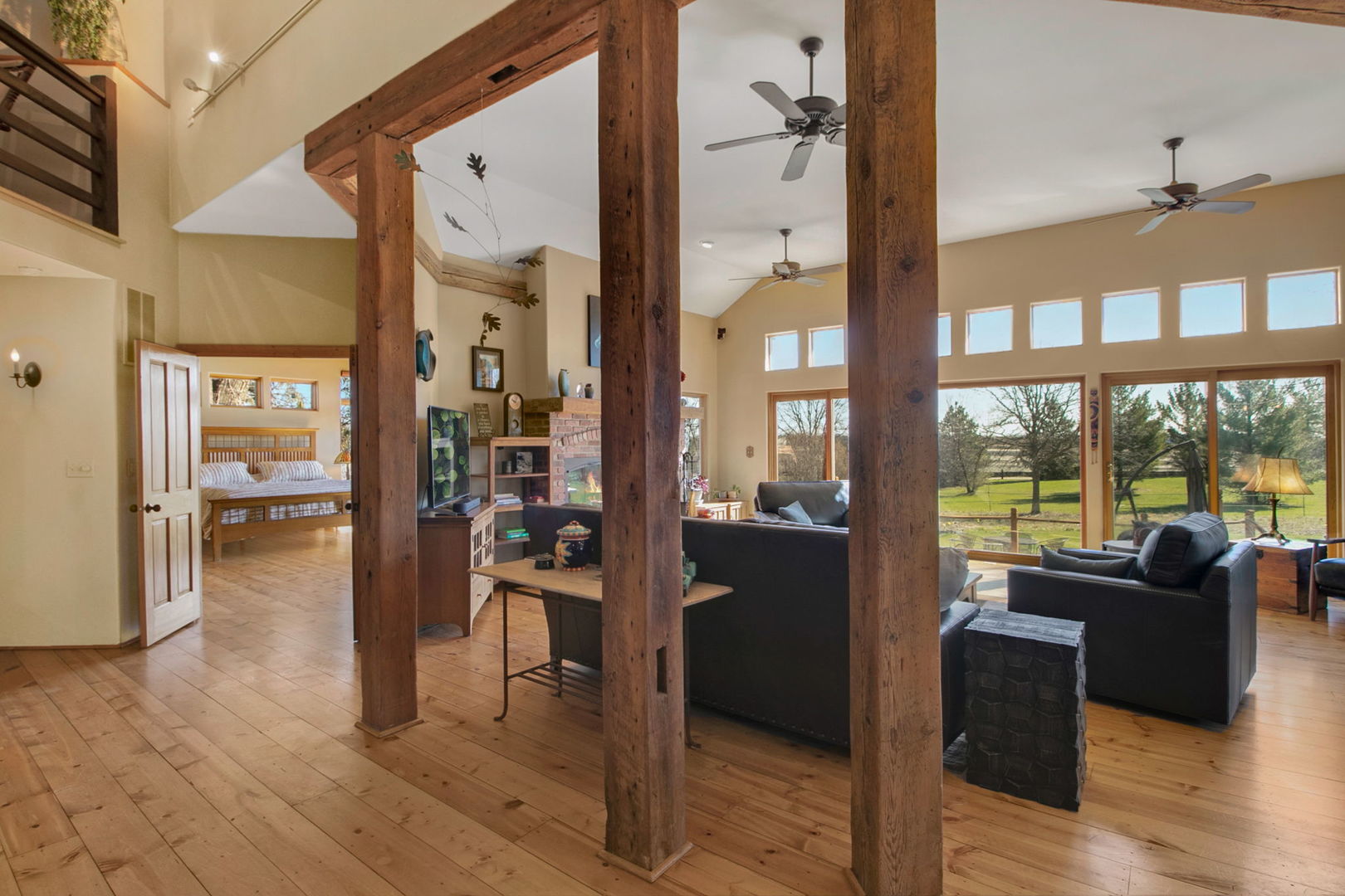 Living room with exposed timber beams, a brick fireplace, and large windows overlooking the yard.